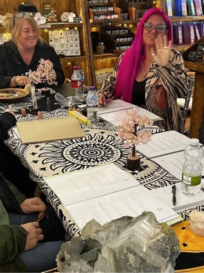Two women sitting at a decorated table with pamphlets, water bottles, and small pink artificial trees. The woman on the right has long pink hair, glasses, and is wearing a leopard print jacket. The woman on the left has blonde hair and is smiling. The setting appears to be a cozy room with wooden shelves and books in the background.