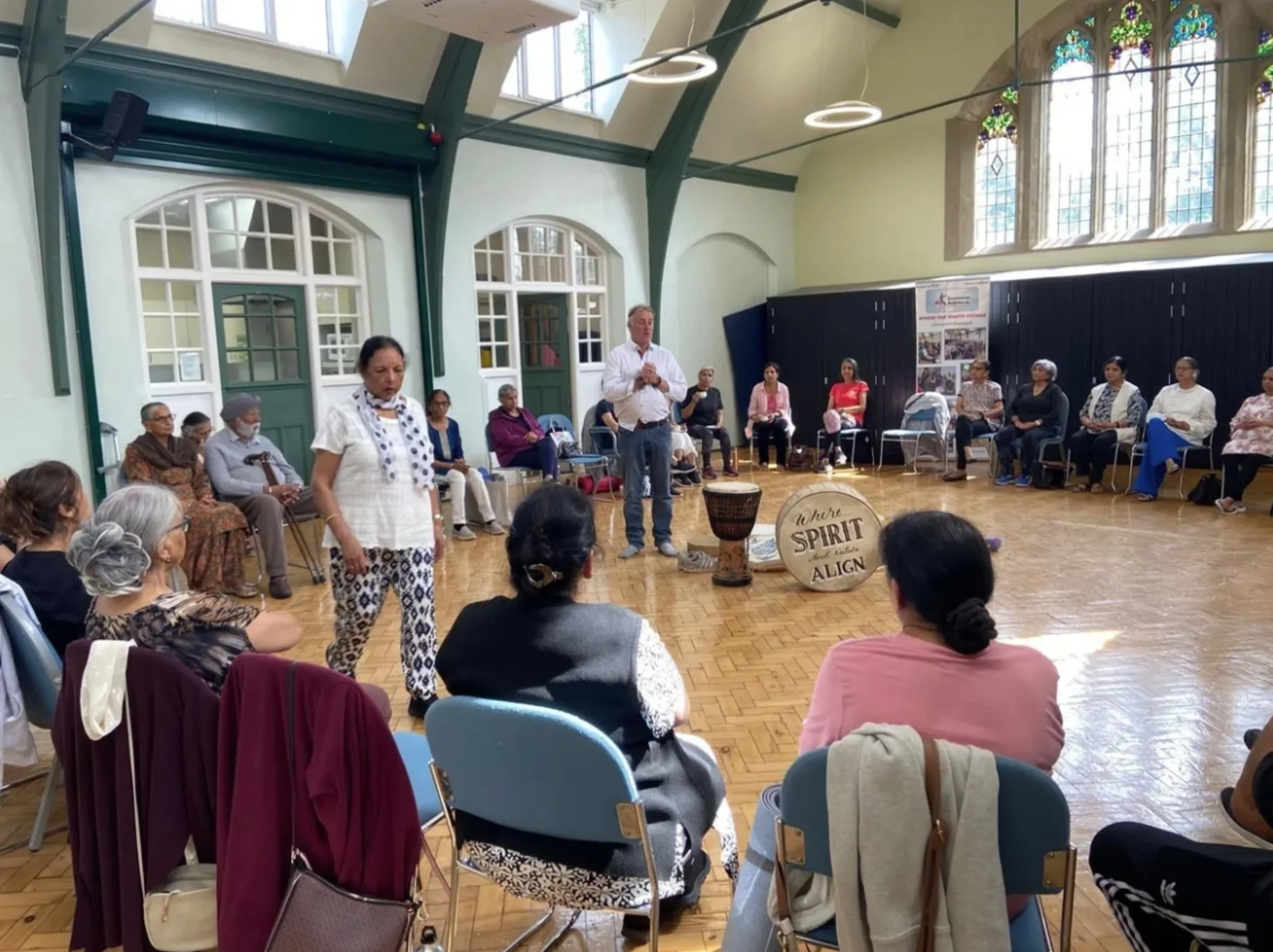 A diverse group of people seated in circle in a large room with tall stained glass windows and high ceiling, participating in a gathering or workshop.