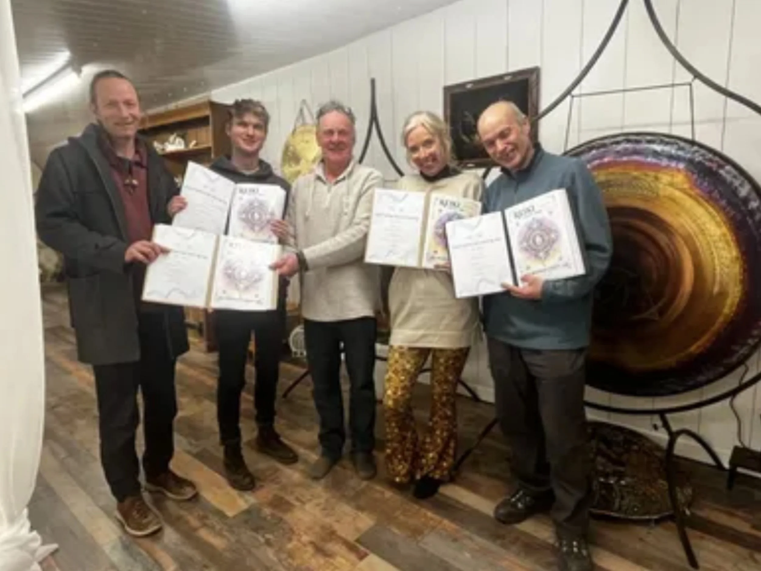 Five people standing indoors, holding certificates, smiling for the camera. One woman is dressed in colorful patterned pants and a beige jacket, three men are in casual attire, and one man is in a dark jacket.