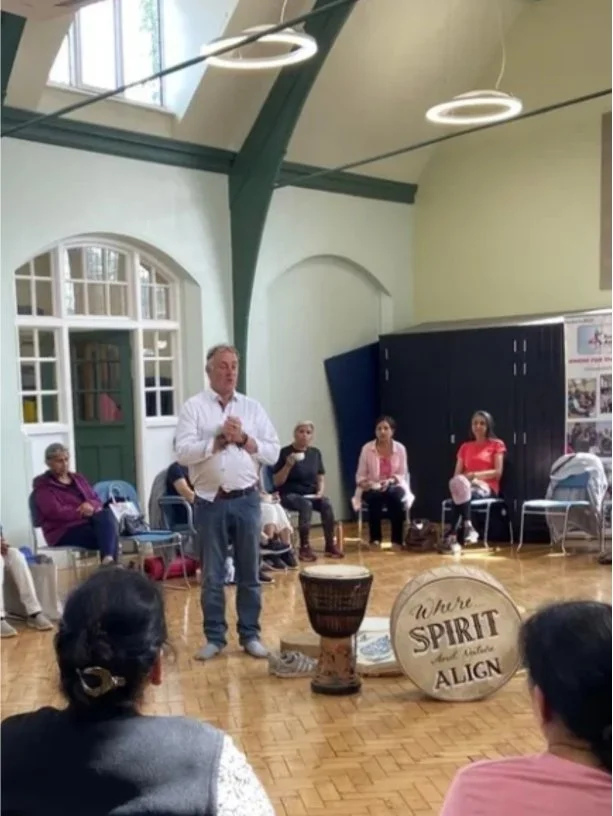 A man in a white shirt and blue jeans speaking to a group of seated people inside a spacious room with high ceilings, large windows, and hanging lights. The group includes diverse women and men, some taking notes or listening attentively. Musical instruments, including drums, are on the floor in front of the speaker, and a large sign reads 'You're SPIRIT and you ALIGN'.