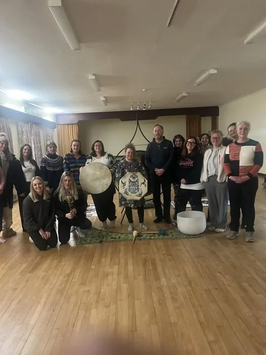 A group of people standing and kneeling in a room with wooden floors and cream-colored curtains. They are posing around large musical instruments including a gong, a drum, a handpan, and a singing bowl.