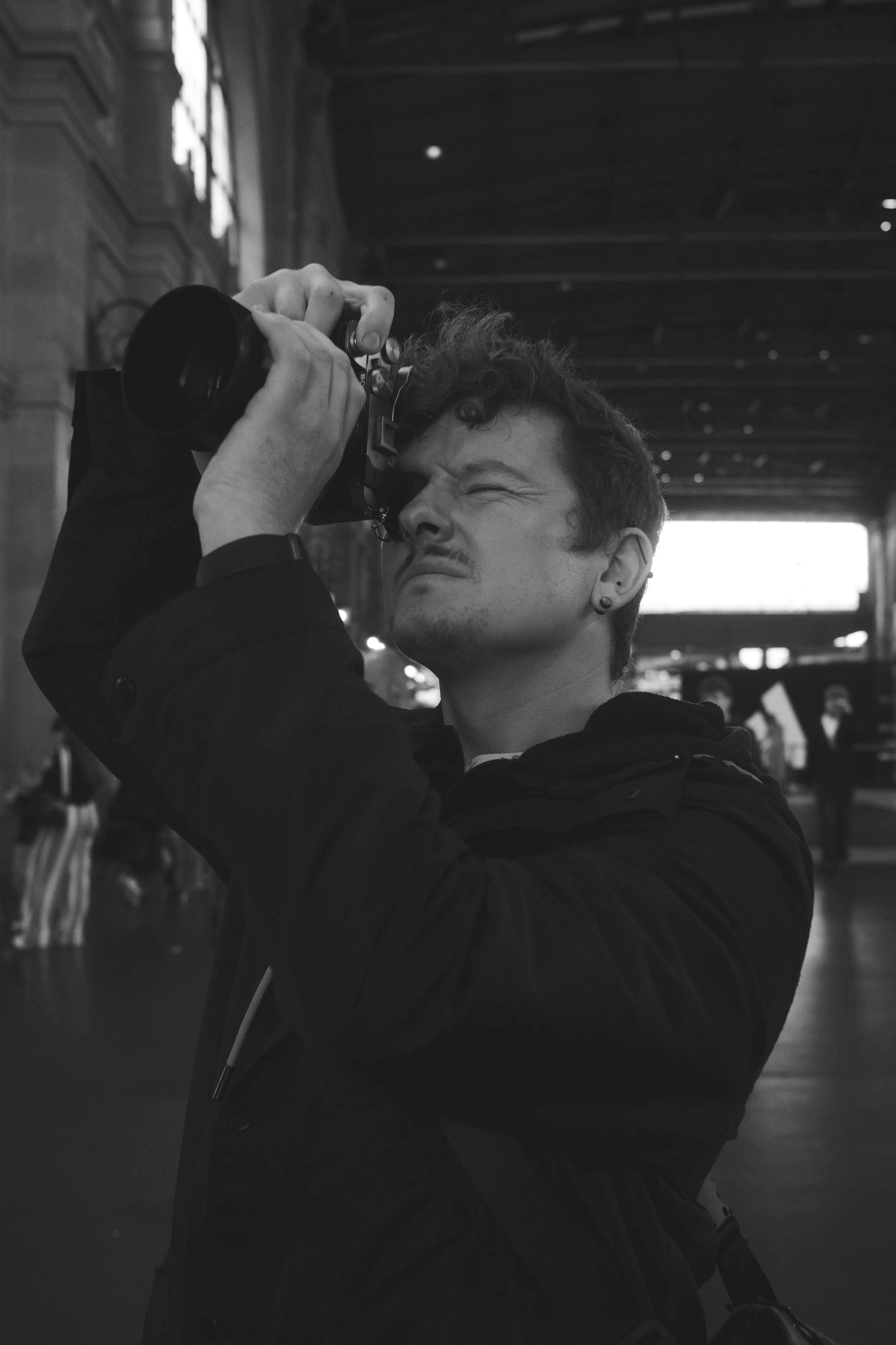 Ein junger Mann fotografiert mit einer Kamera in einer großen Halle, die im Schwarz-Weiß-Stil aufgenommen wurde.