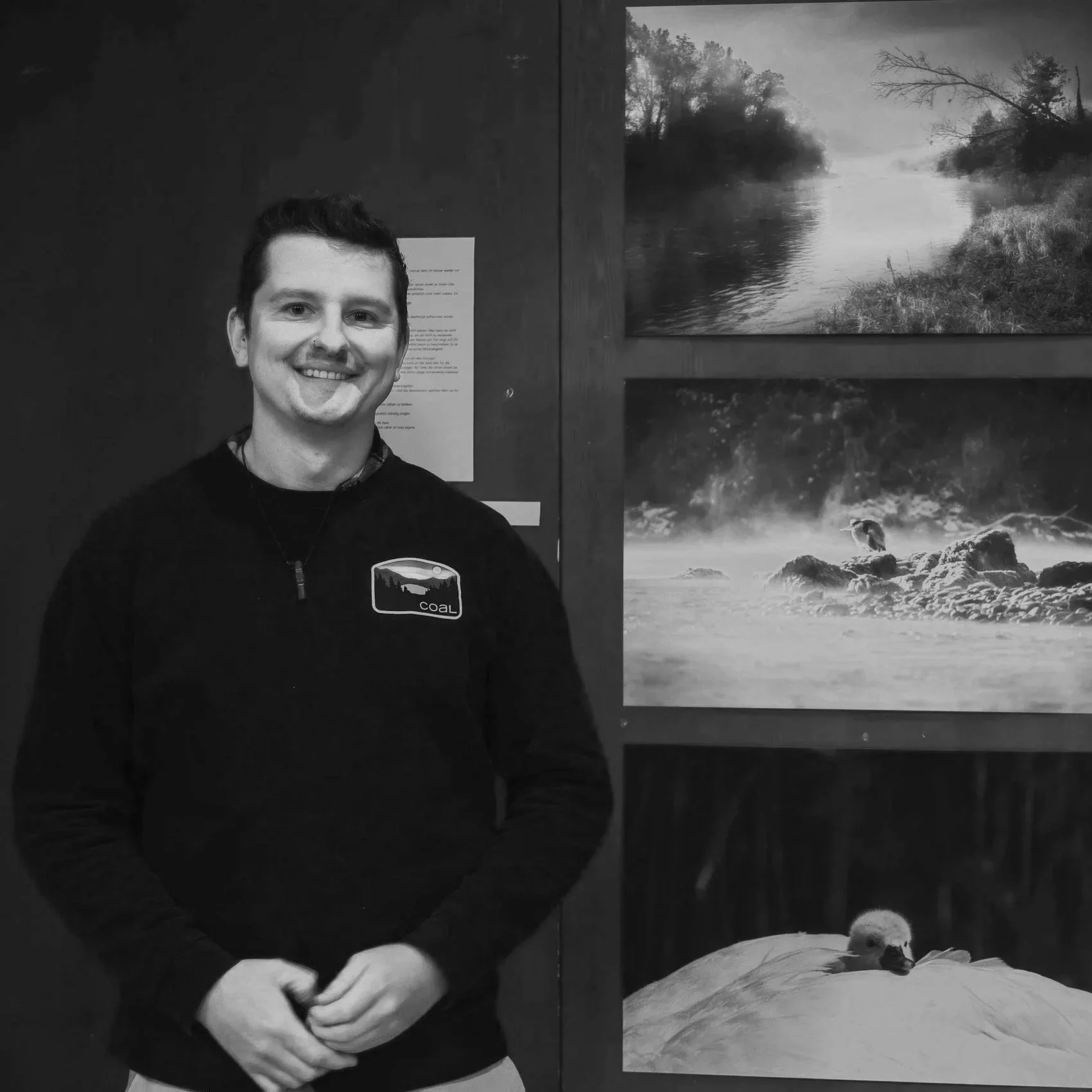 Ein lachender junger Mann steht vor einer Wand mit drei Tier- und Landschaftsfotografien. Er trägt einen schwarzen Pullover mit einem Logo, das 'COAL' heißt.