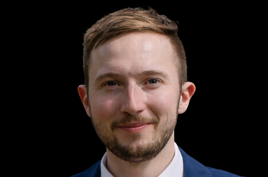A young man with light brown hair, a beard, and blue eyes smiling, wearing a navy blazer and a white shirt, against a black background.