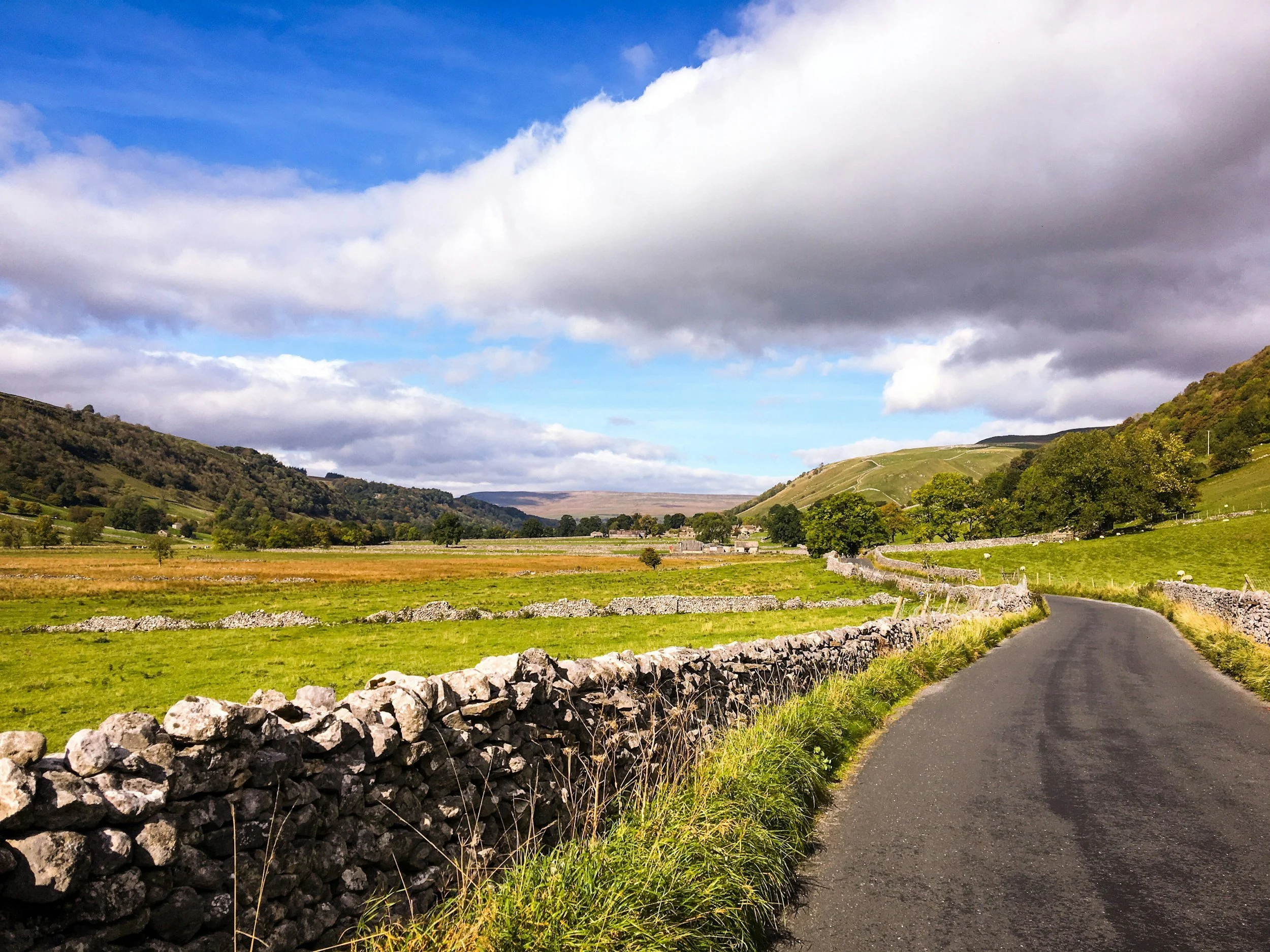 A winding narrow country road passes through green fields with stone walls and is surrounded by rolling hills and mountains under a partly cloudy sky.