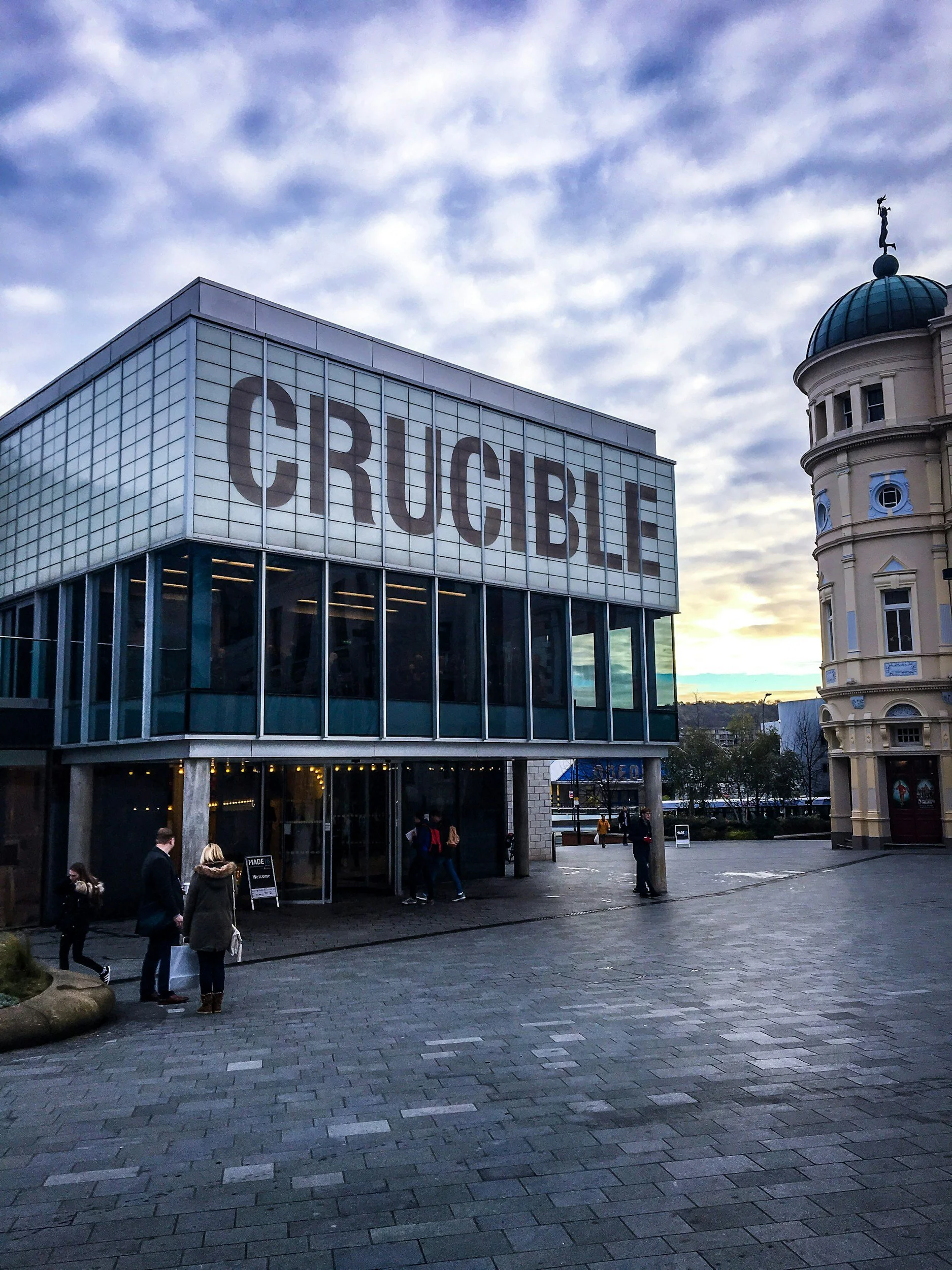 A modern building with large glass windows and a sign that reads 'CRUCIBLE' in big letters. There are a few people walking and standing outside, and a historical building with a dome is visible to the right. The sky is partly cloudy, and the scene appears to be during dusk or early evening.