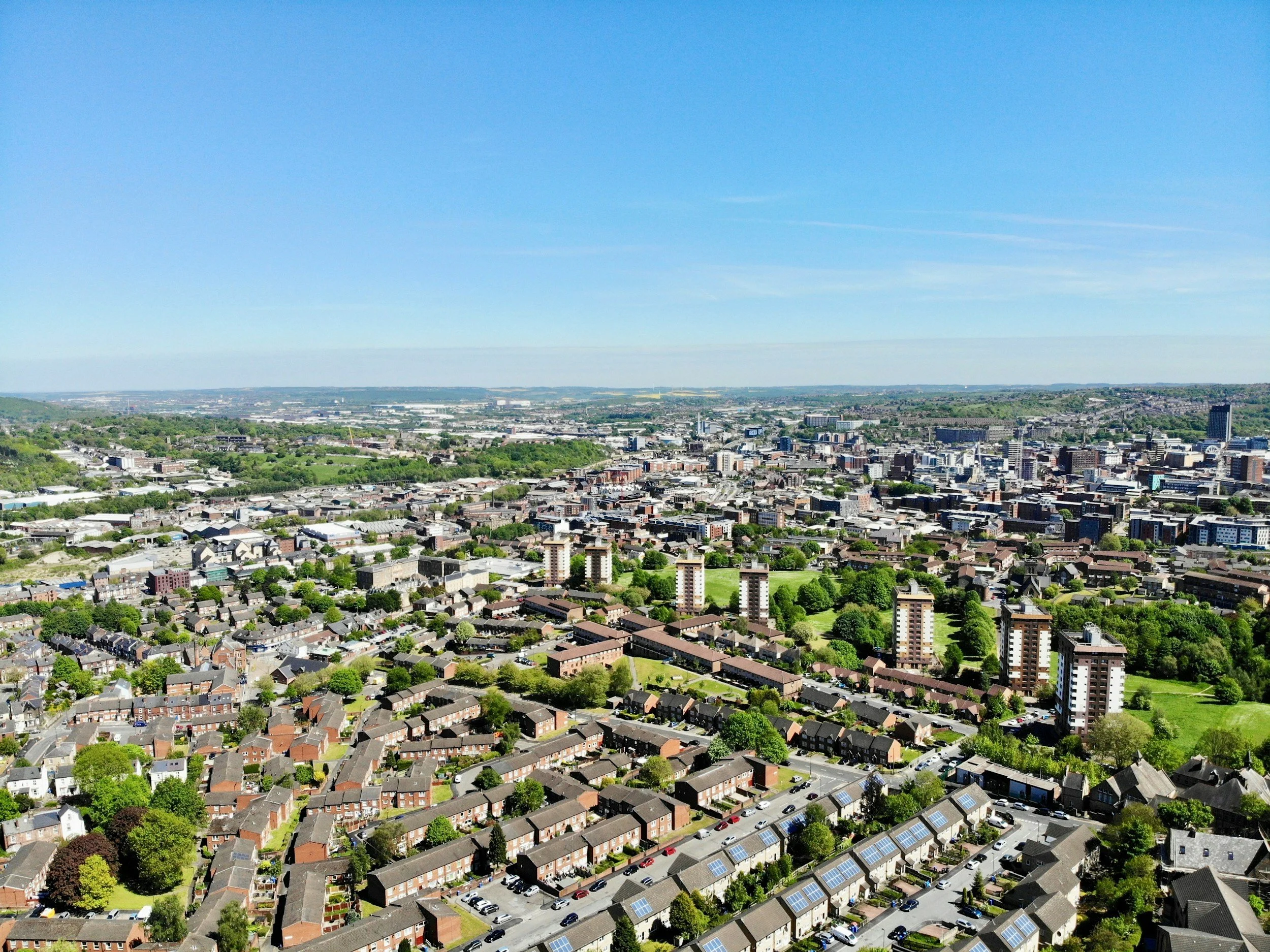 Aerial view of a city with a mix of residential houses, high-rise buildings, green parks, and a distant horizon under a clear blue sky.