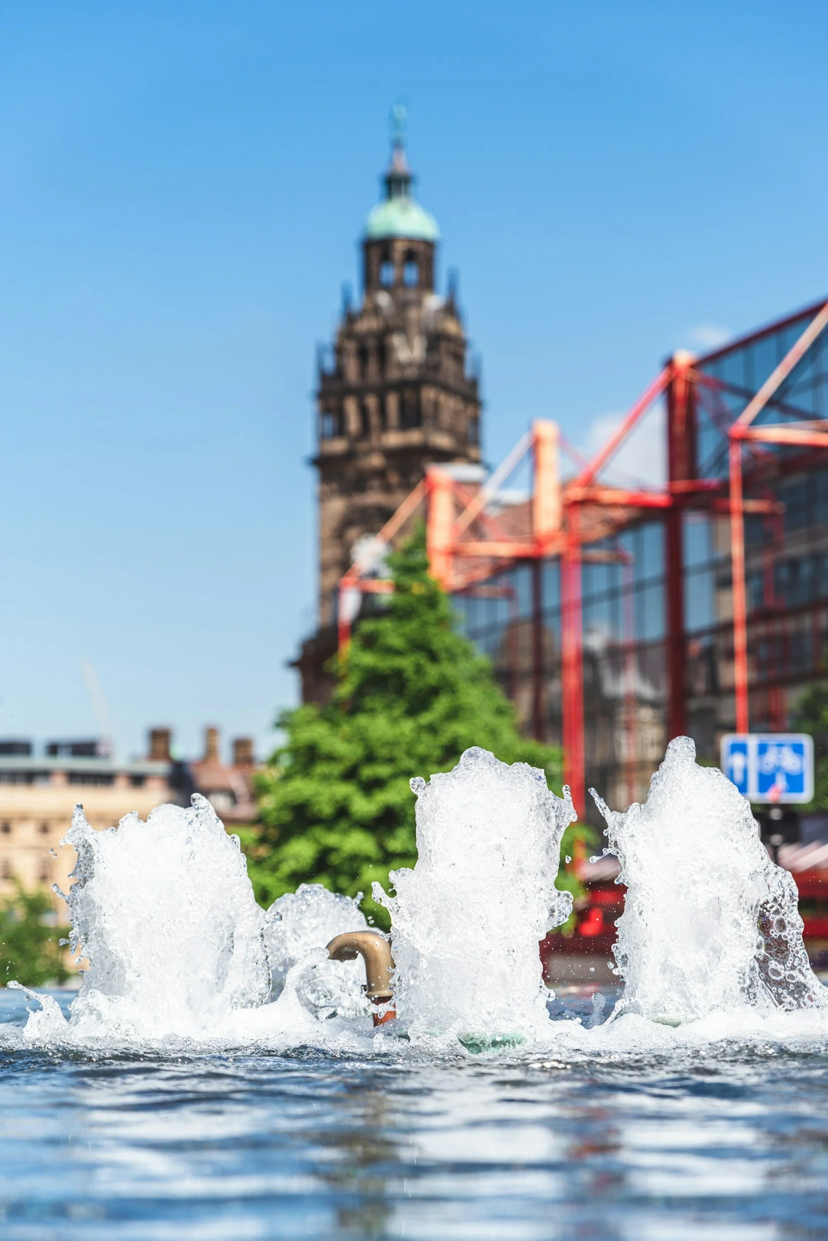 Fountain with water splashing in the foreground, historic clock tower, and modern glass building with red frame in the background under a clear blue sky.