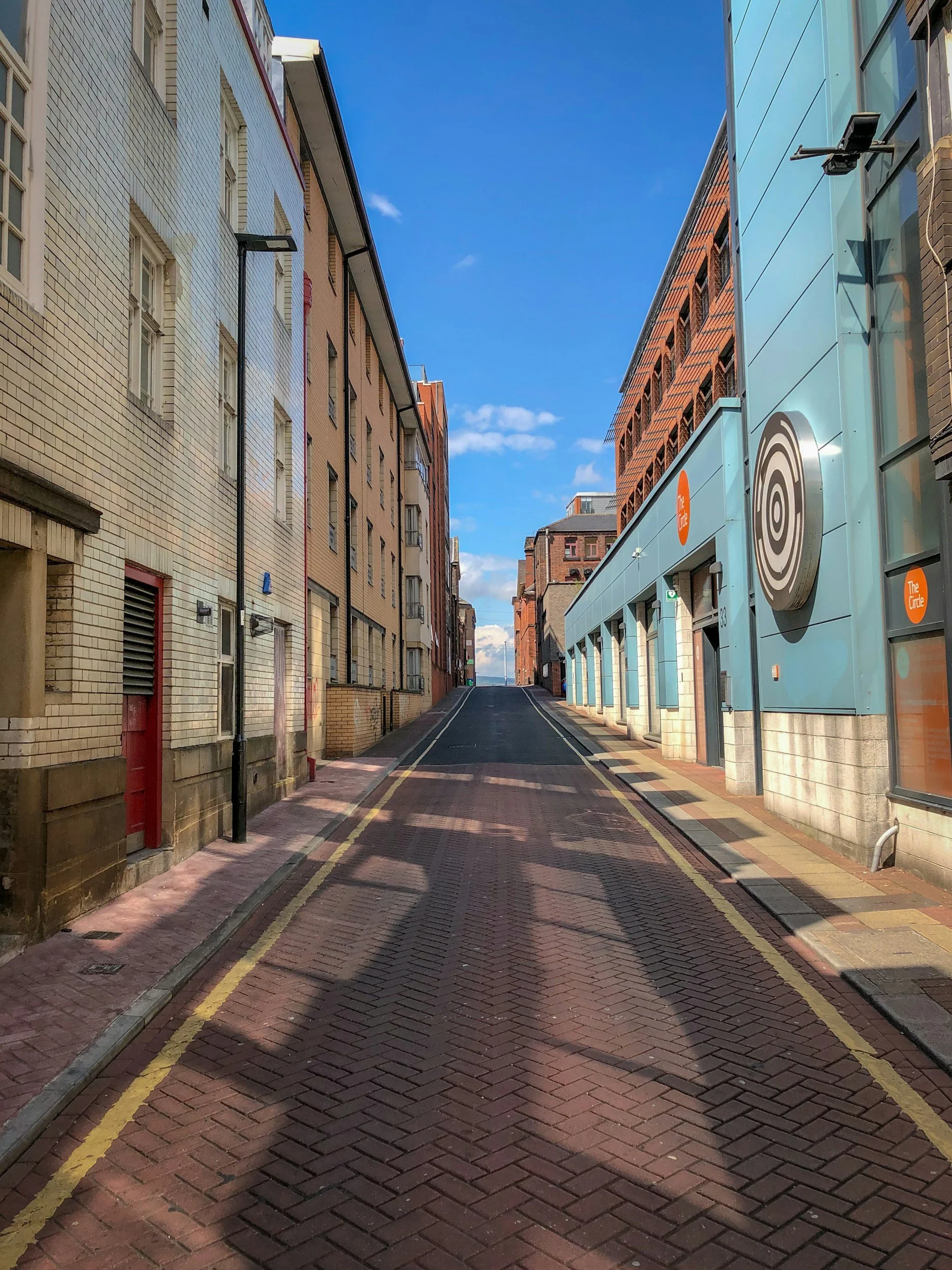 Empty city street with brick pavement, lined with tall buildings on both sides under a blue sky with some clouds.