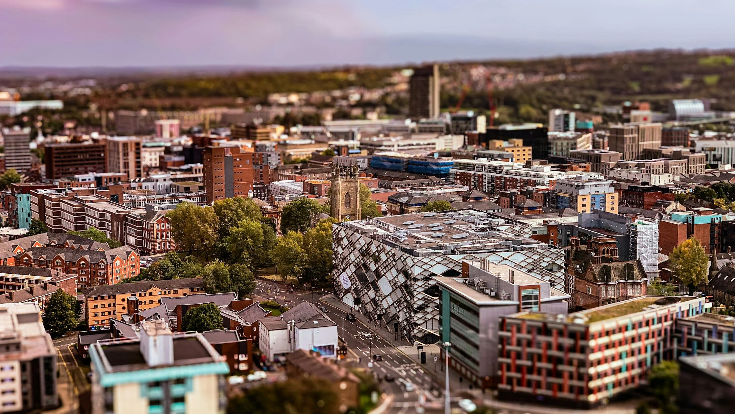 Aerial view of a city with a mix of modern and historic buildings, with tree-lined streets and a church tower in the center, under a partly cloudy sky.