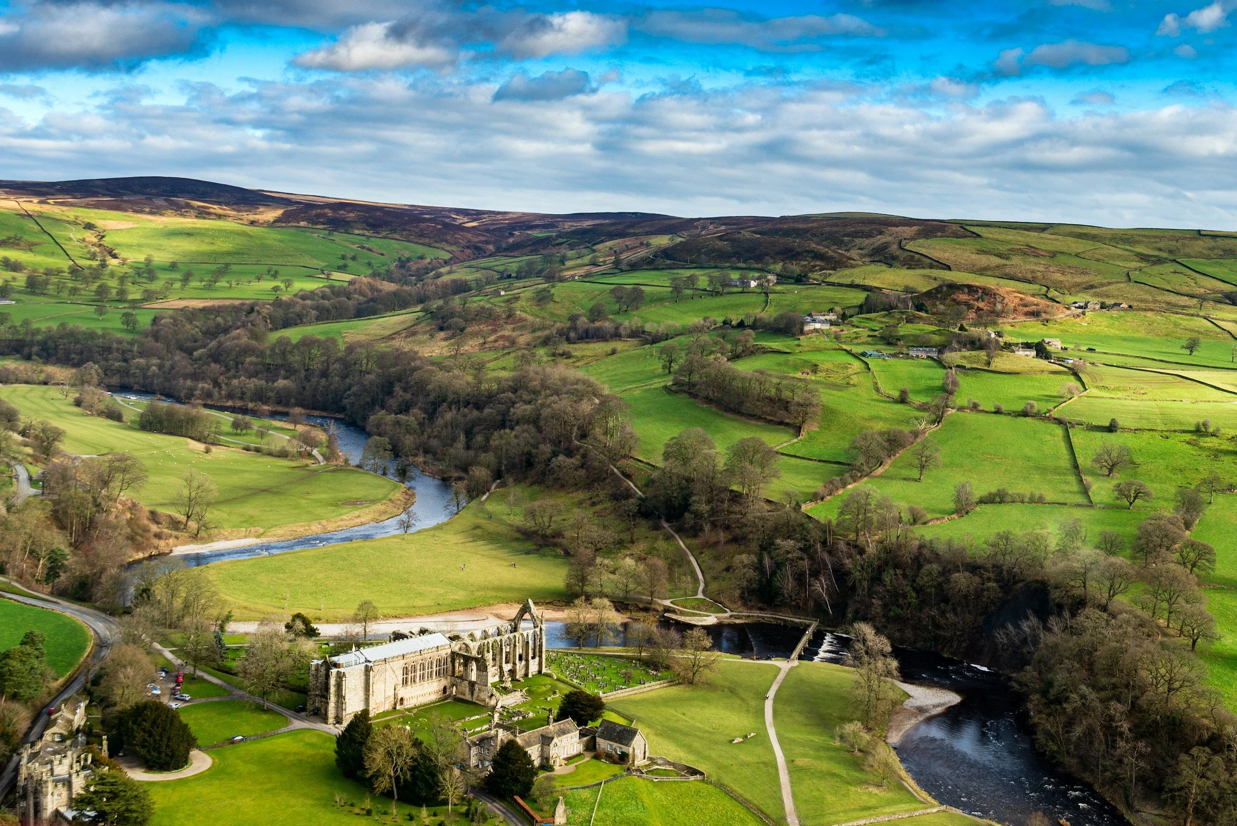 Scenic landscape of rolling green hills, a winding river, and a historic stone building, with a partly cloudy sky overhead.