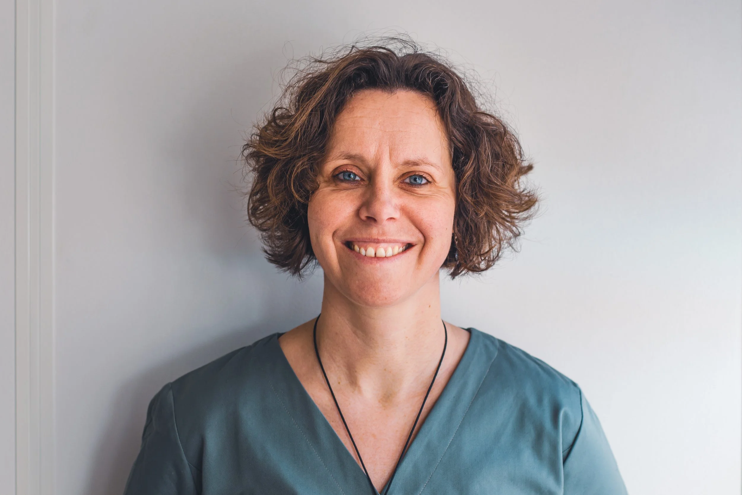 Une femme souriante avec des cheveux bouclés bruns, portant un haut d'un vert foncé, se tenant devant une paroi blanche.