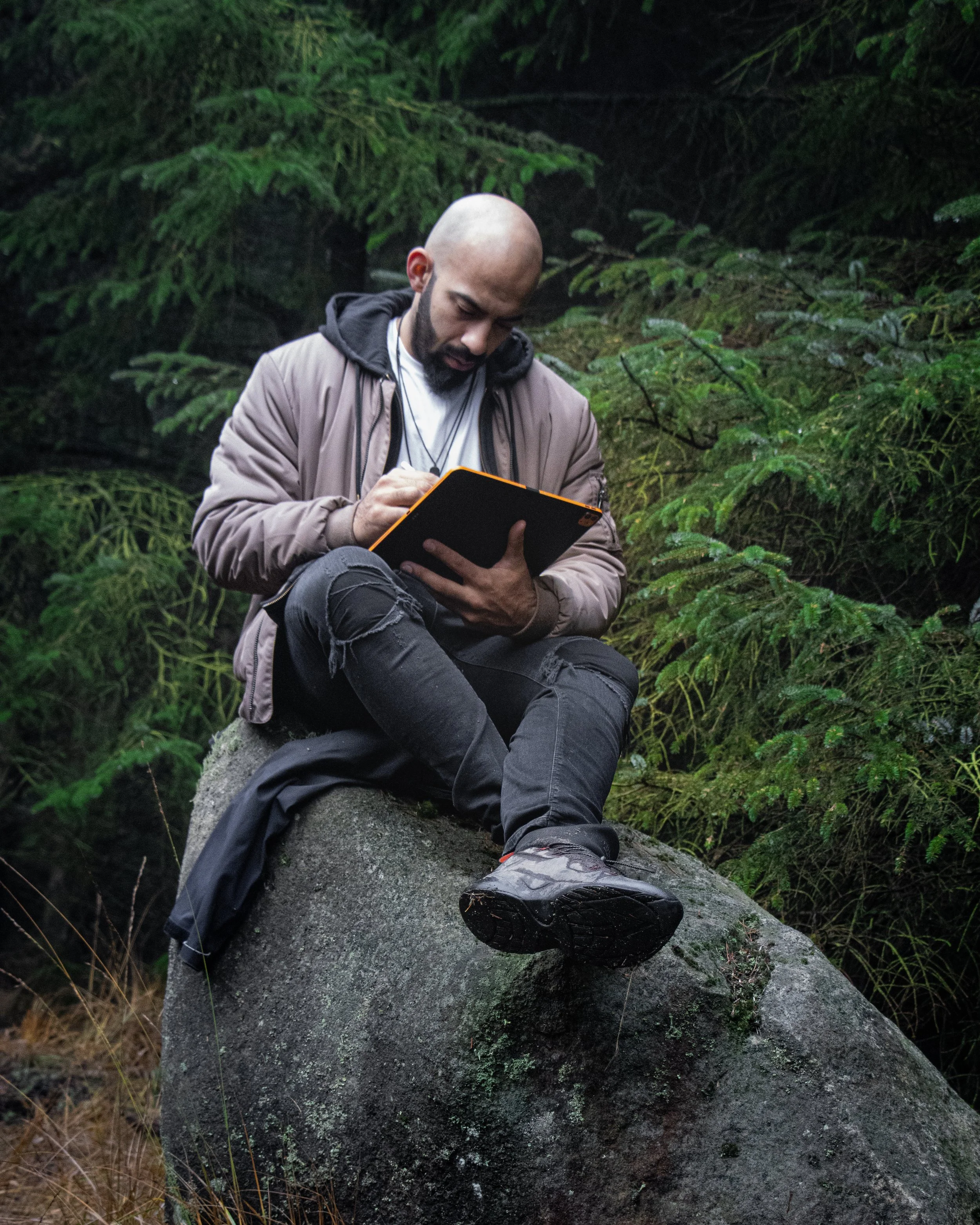 Man sitting on a large rock in a forest, writing or drawing in a notebook.