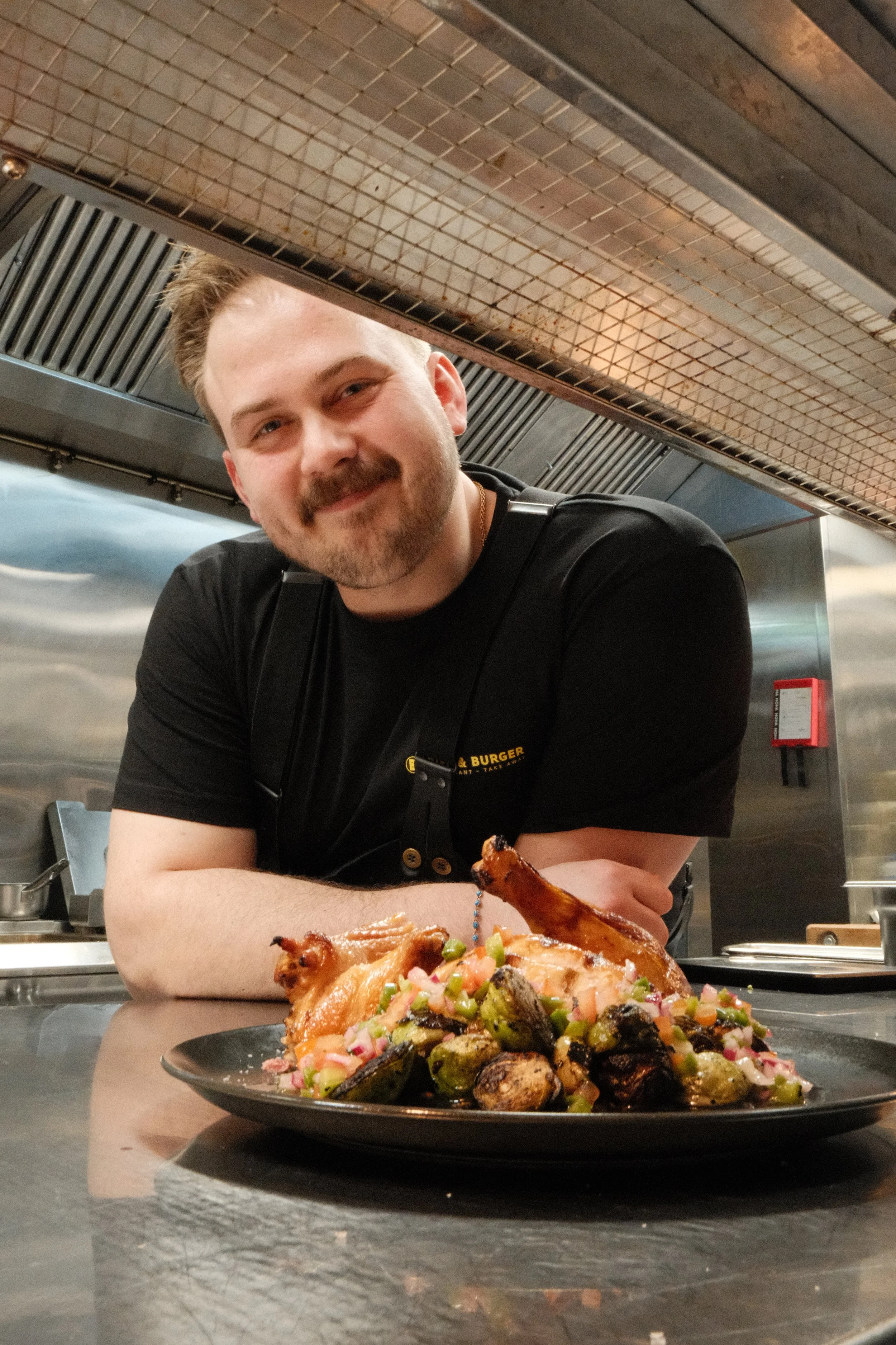 En mann smiler foran en tallerken med mat, inkludert kylling og grønnsaker, i et kjøkken.