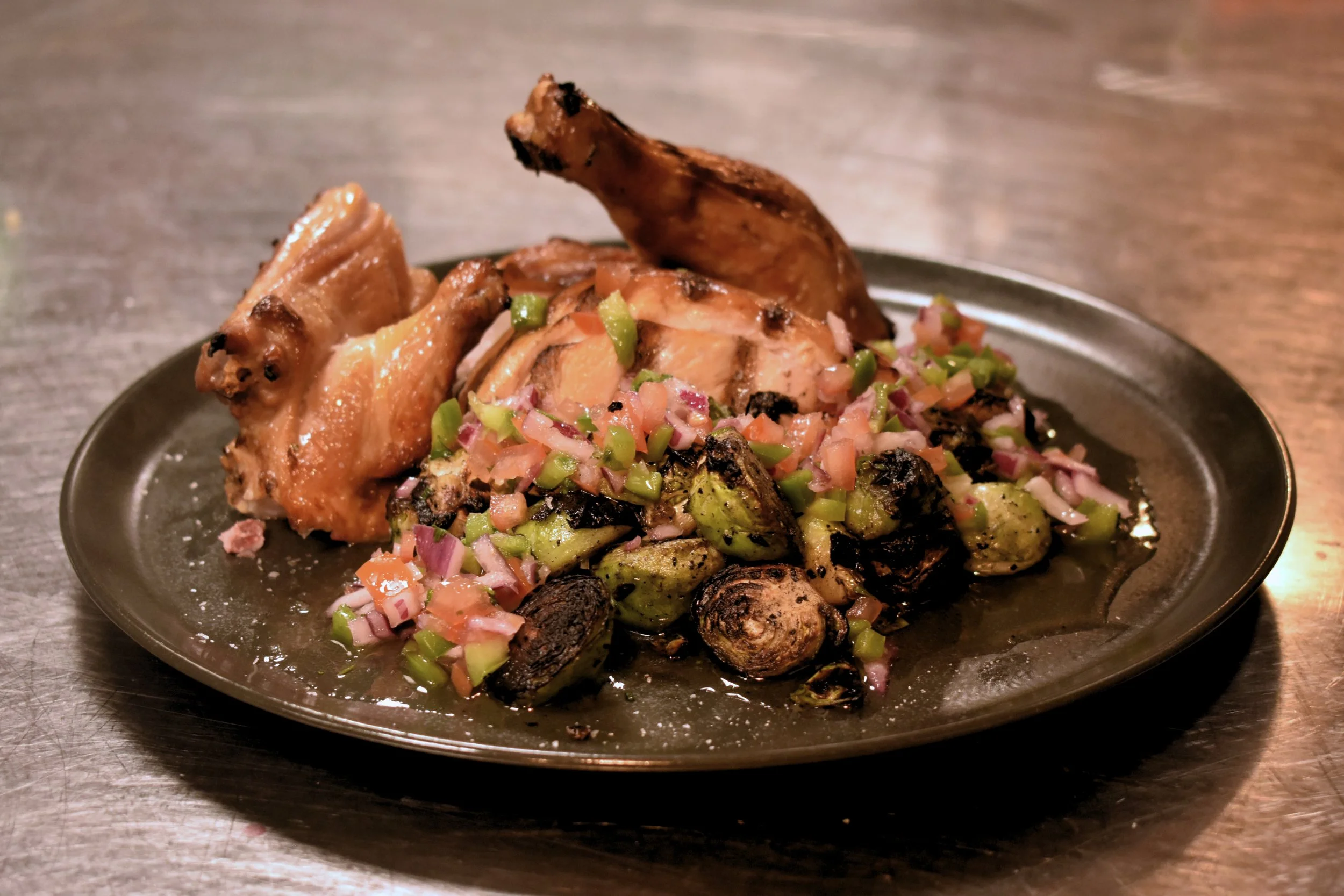 Grillet kyllingestykke med en siderett av grillede tomater, hakkede løk, og grønne pepperstykker på en svart oval tallerken.