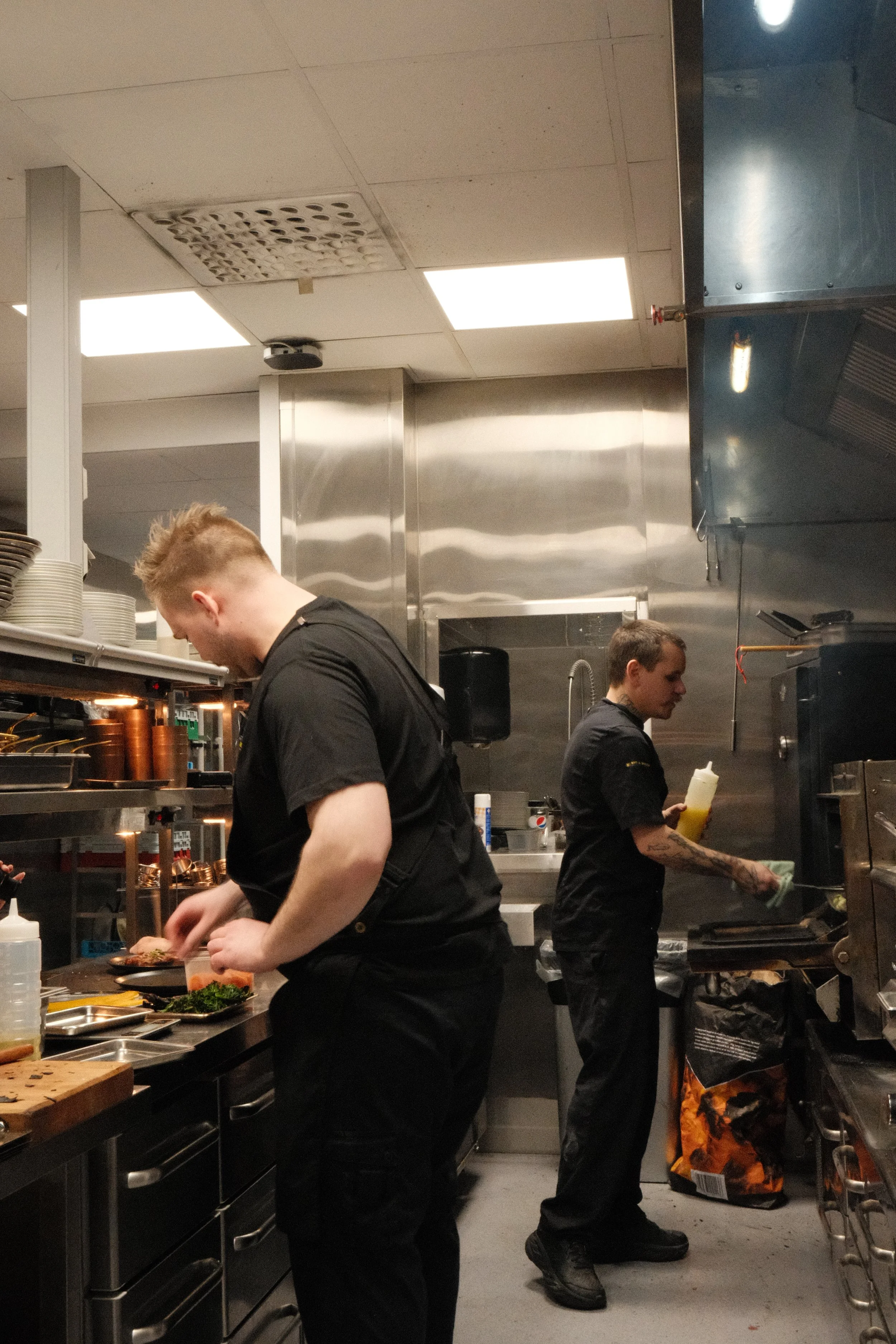 To kokker forbereder mat i en kommersielt kjøkken, med metallvegg, kjøkkenutstyr og krydder rundt seg.