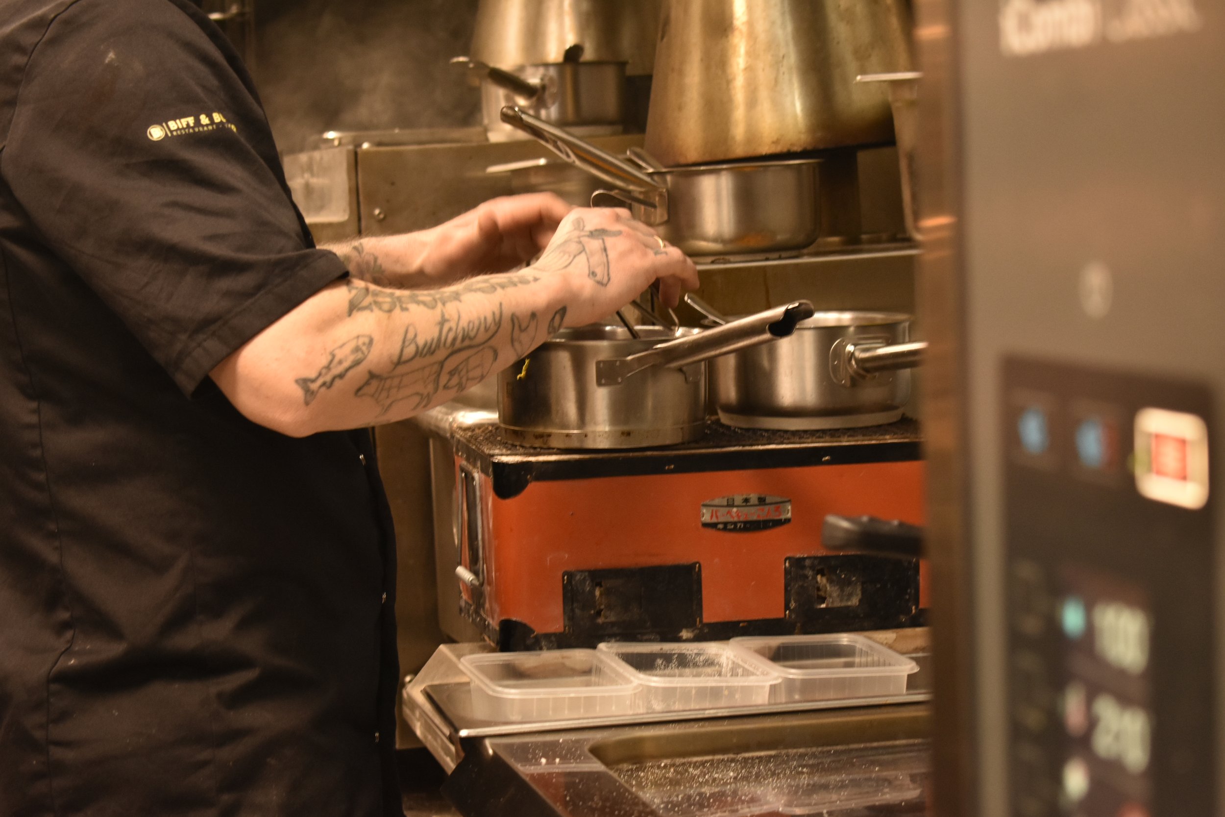 En person med tatoveringer på armen lager mat på komfyr i et kjøkken.