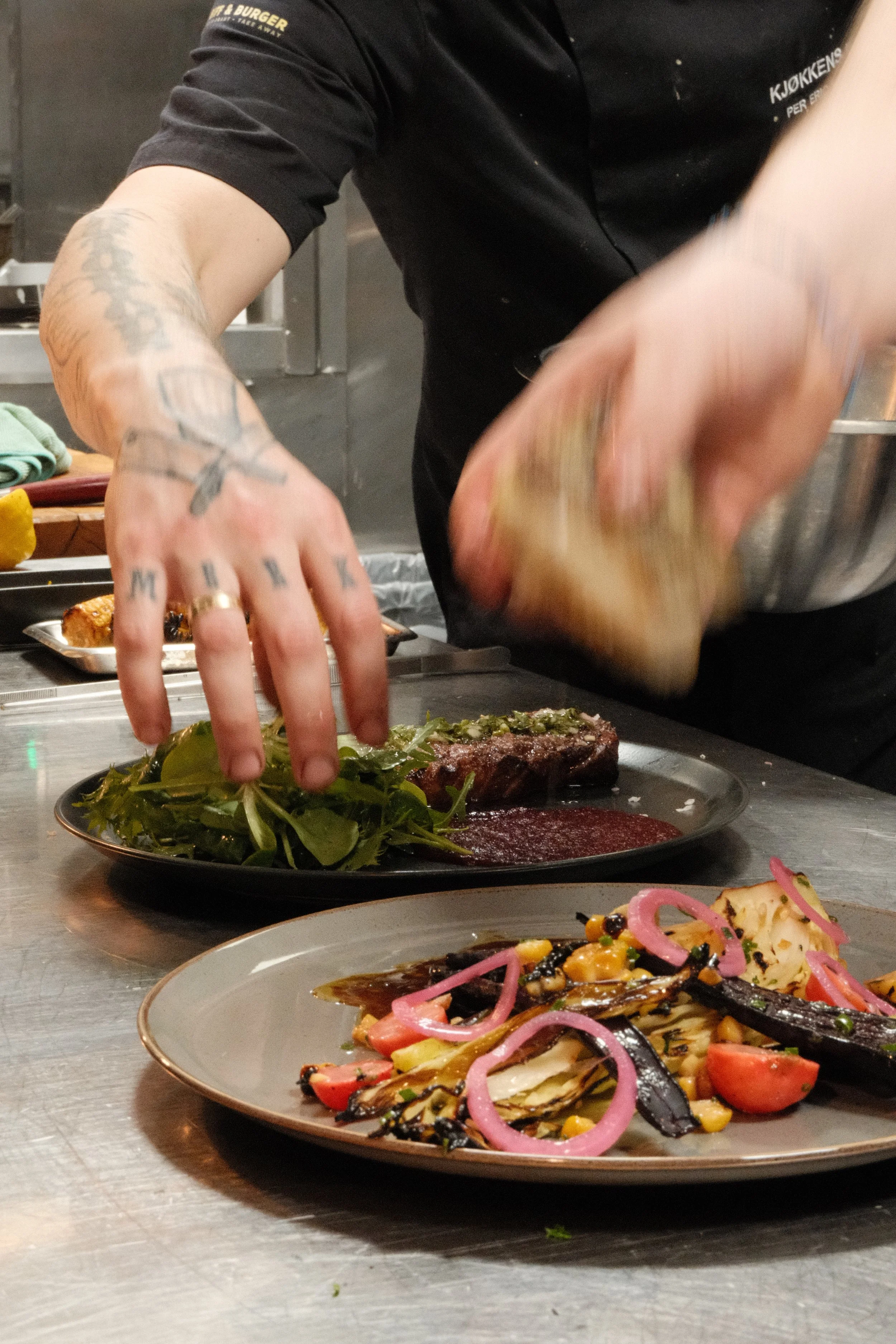 En kok legger opp en tallerken med salat og en annen tallerken med grillmat, inkludert auberginer, tomater og løk, på en stålkjøkkenbenk.