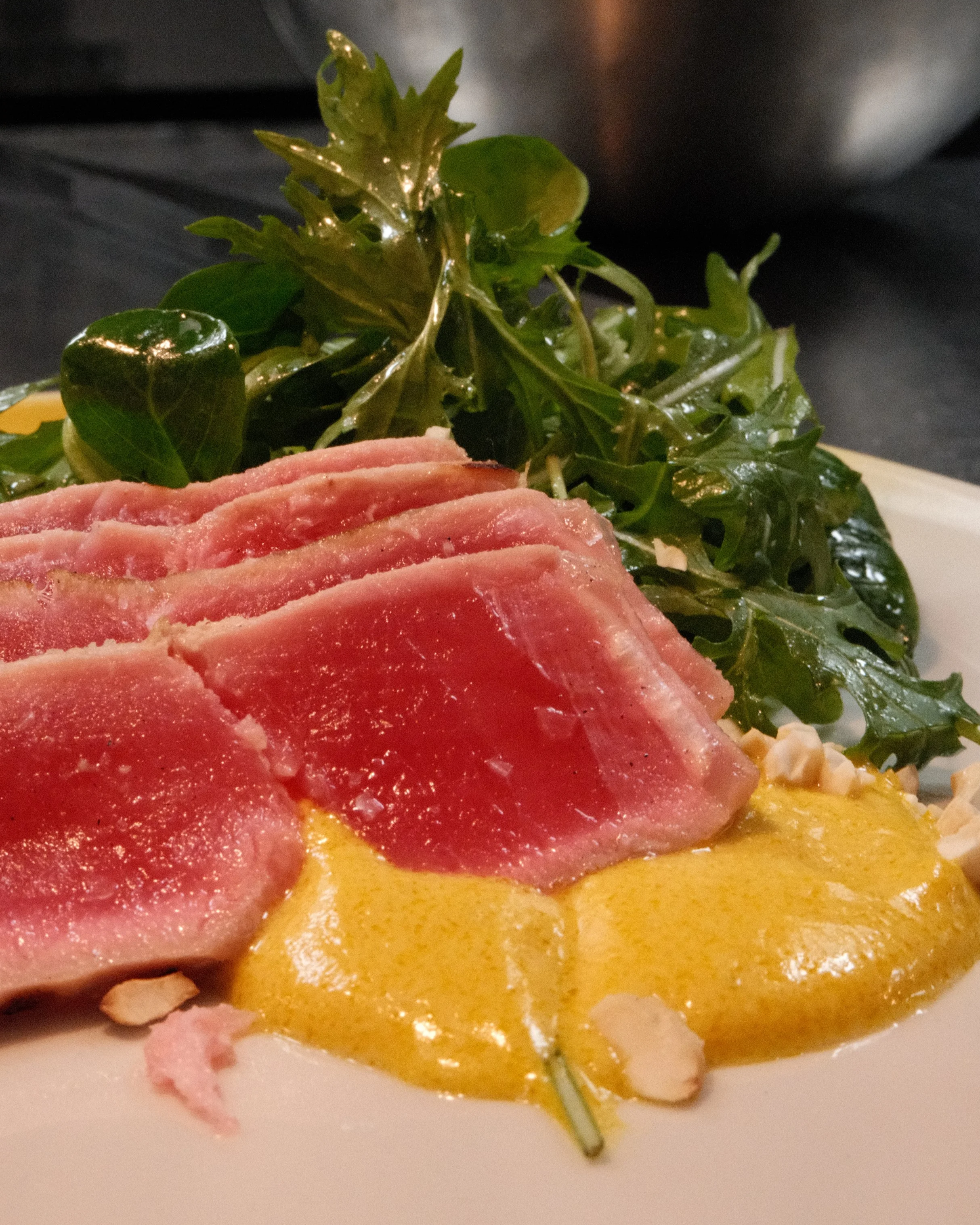 Tykke skiver av rå tunfisk på en tallerken med en gul saus, grønn salat og noen mandler.