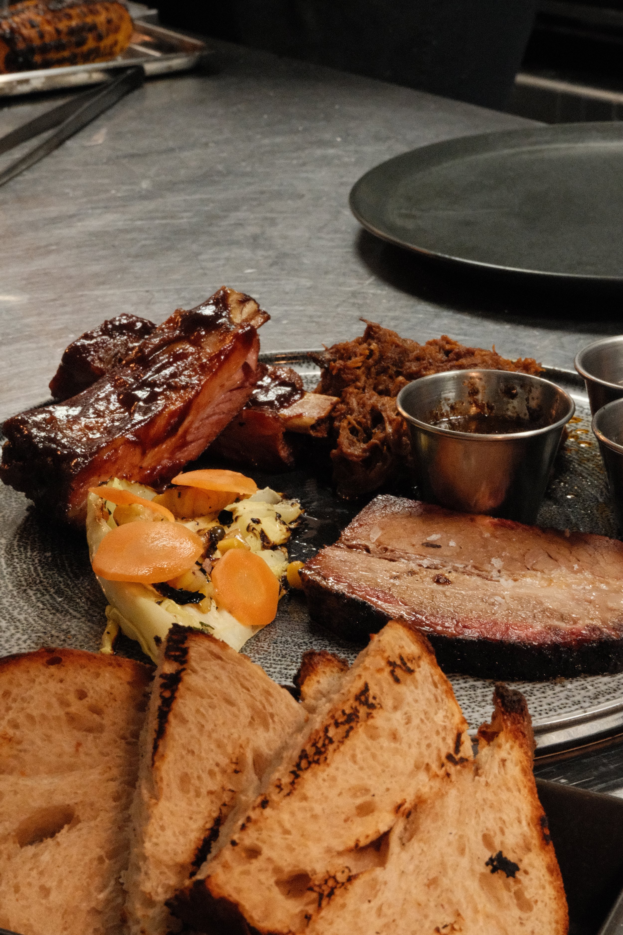 Et fat med grillet kjøtt, potetsalat, brød og små kopper med saus.