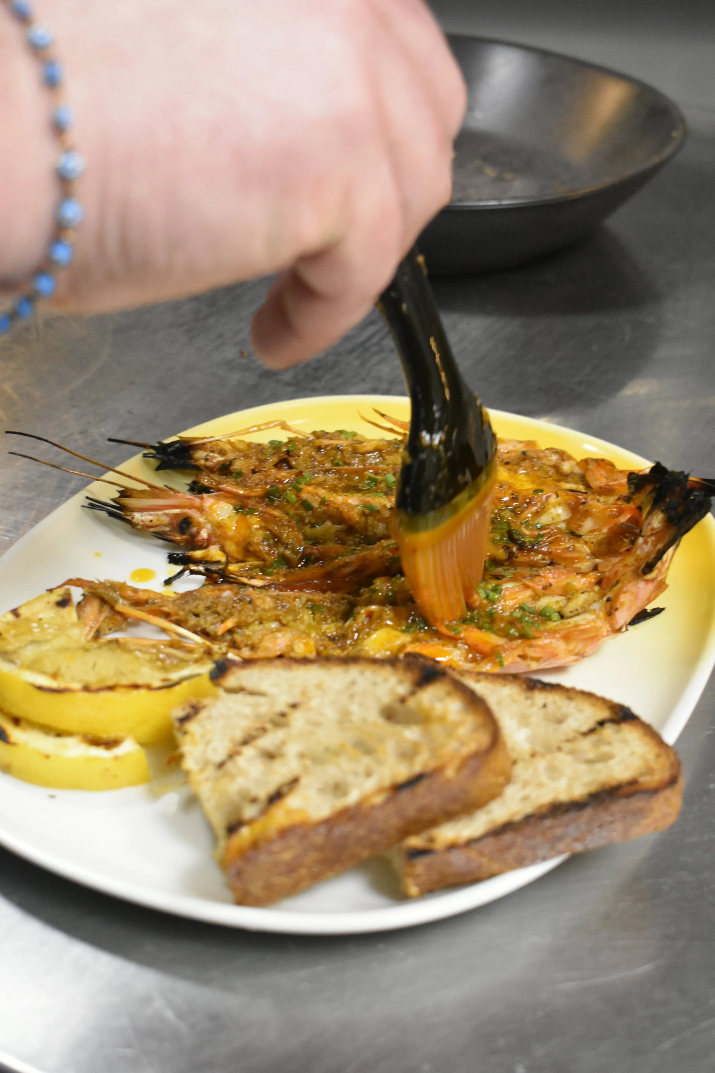 En person legger sausje over grillet reker på en hvit tallerken med brød og sitronbåter, på et stålbord.