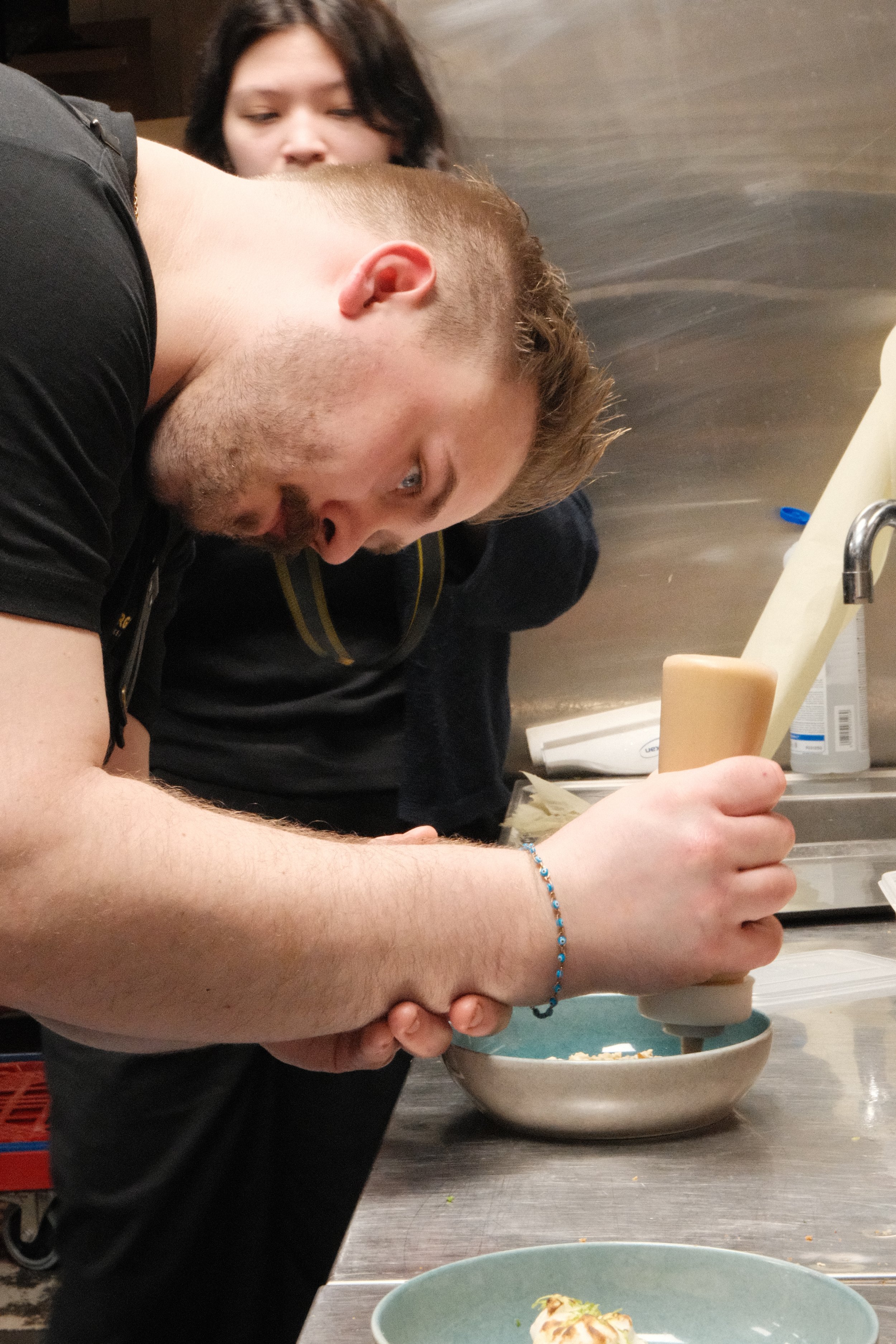 Person som bruker kjøkkenutstyr, med en annen person i bakgrunnen.