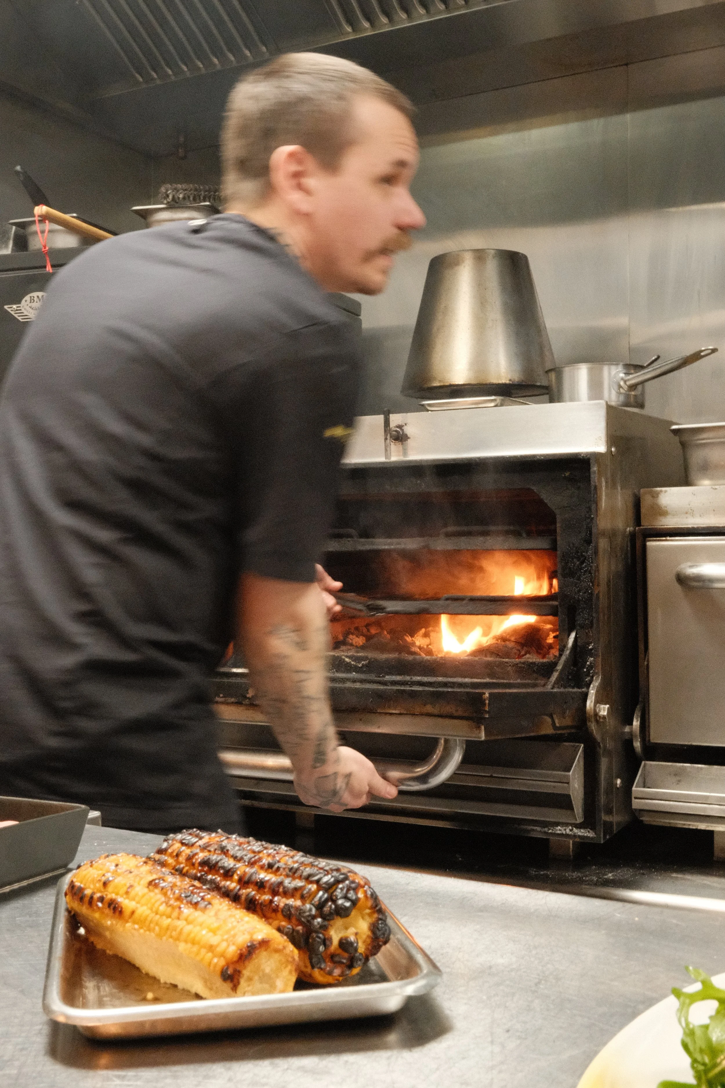 En kok som fyller opp en grill med kull, med to grillede maiskolber på en metallfat foran ham.