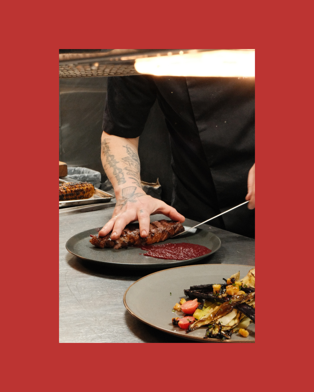 En kok som skjærer og anretter en stor, grillet kjøttbiff på en svart tallerken, med en rød saus ved siden av. På bordet er også en tallerken med grillet grønnsaker.