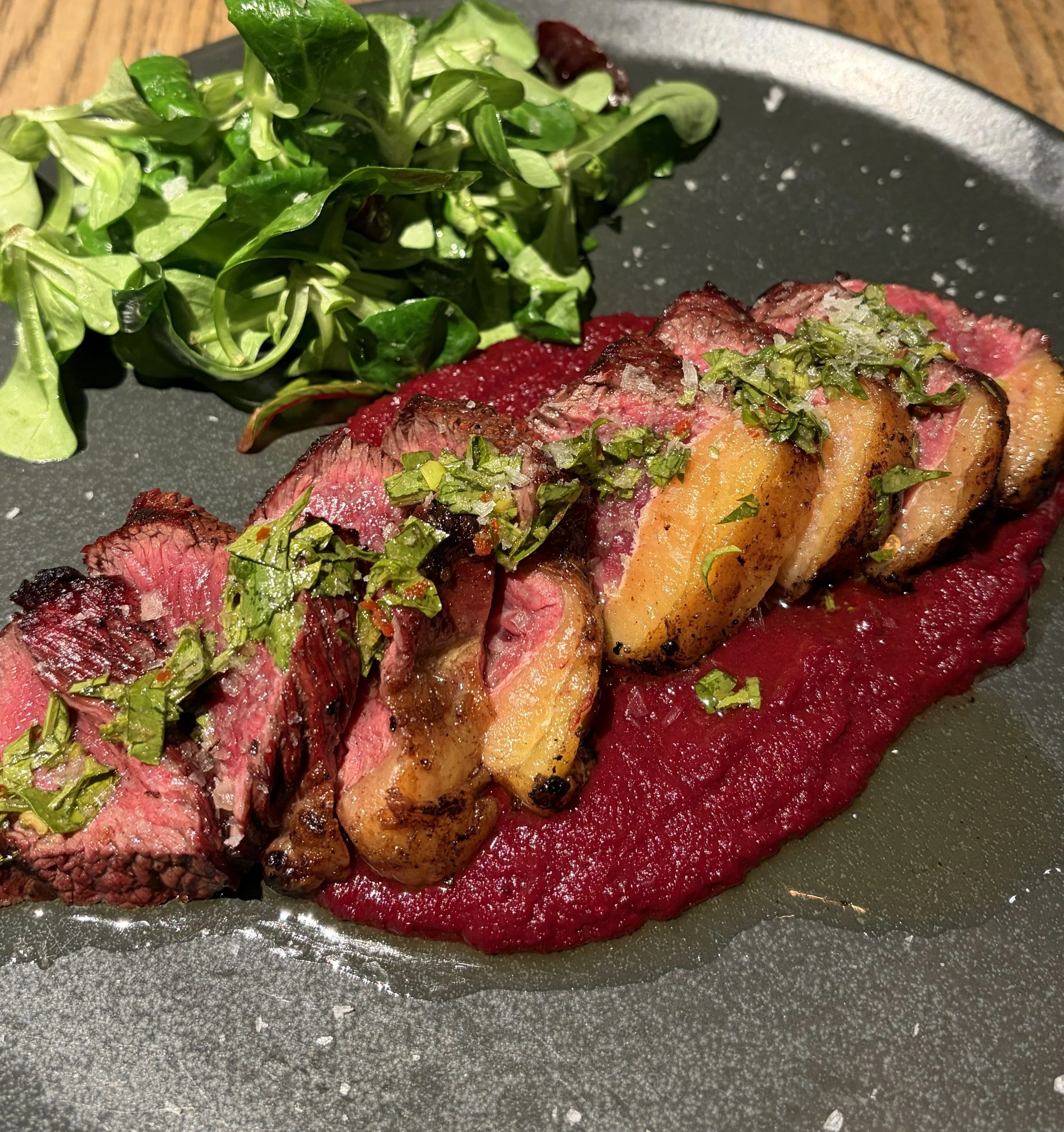Biffstykker med grønn urtesaus, servert på en mørk tallerken med rødbetpuré og en liten grønn salat.