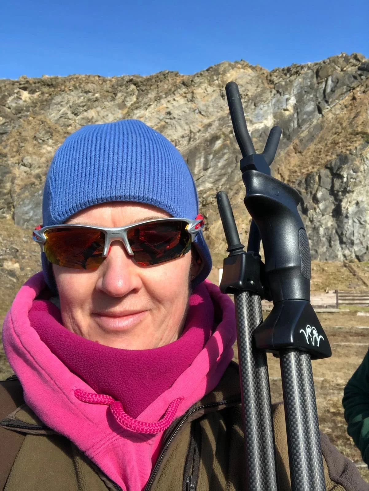 A woman stands facing the camera. She is wearing a blue beanie hat, wraparound sunglasses and a pink hoodie. SHe is holding a pair of Blaser shooting sticks and standing outdoors on a rifle range