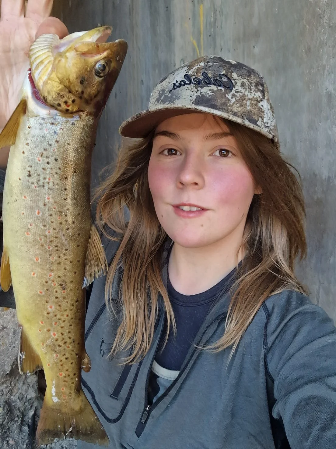 Close-up of a smiling young woman wearing a camouflage Cabela's baseball cap and an outdoor jacket in a natural setting, whilst holding a trout she has fished for and caught up to the camera