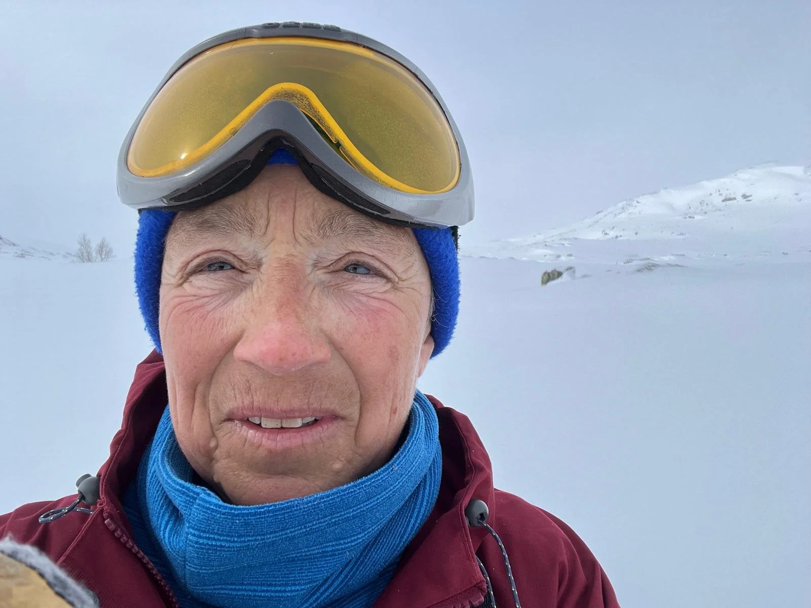 A woman stands outdoors wearing orange tinted ski goggles, a blue hat and neck scarf, and a red jacket, in a polar landscape