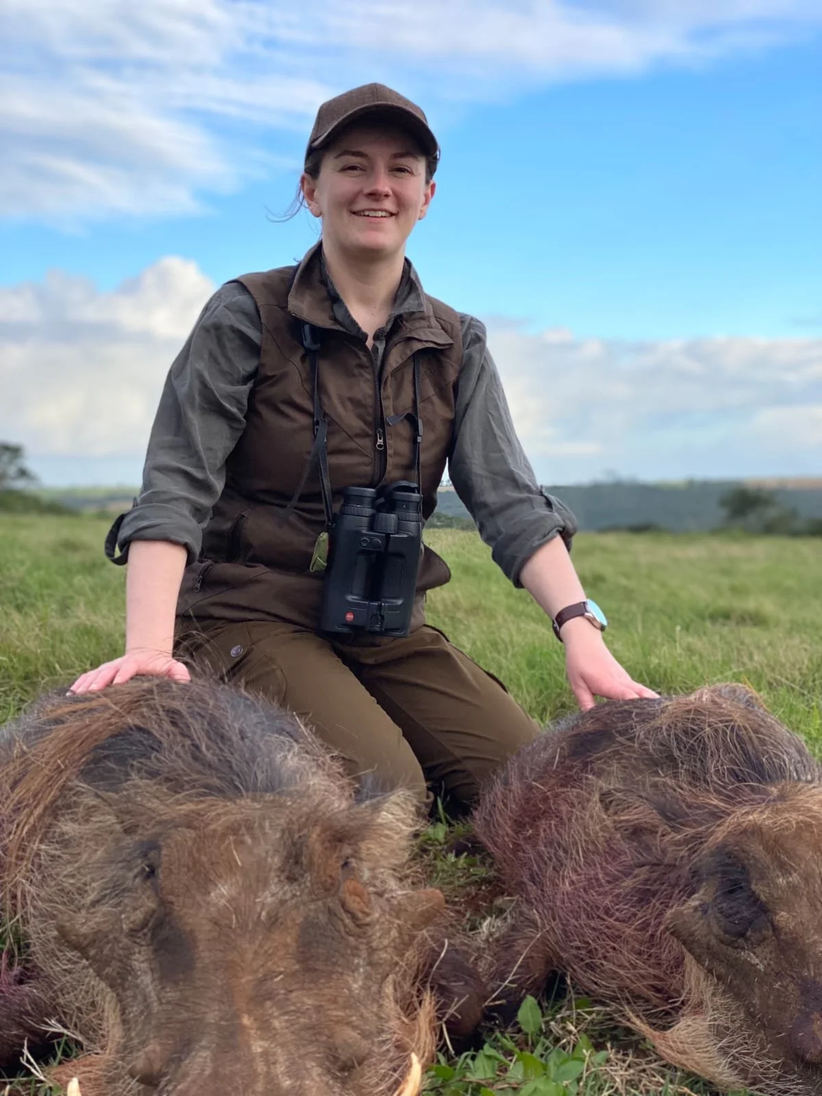 A woman smiles at the camera whilst kneeling outdoors in a grass area in Africa. She is wearing a brown baseball cap, a brown Harkila waistcoat and a dark green shirt. She has a pair of Leica binoculars around her neck