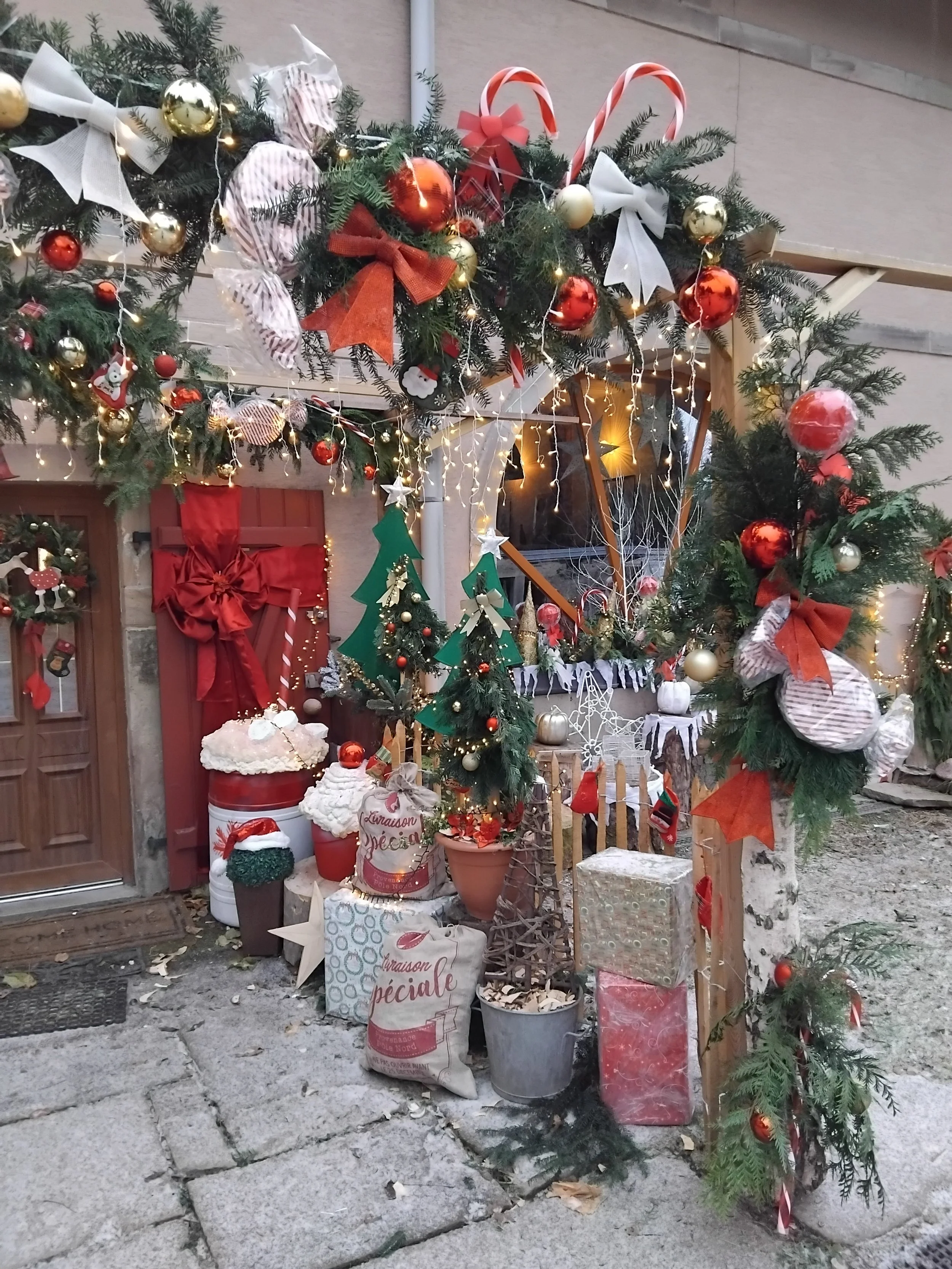 Décoration de Noël avec guirlandes, boules, rubans, sapins miniatures, cadeaux et lumières installée devant une maison.
