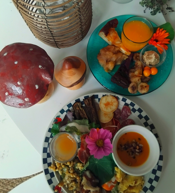 Petit déjeuner avec des plats salés et sucrés, un verre de jus d'orange, un champignon, une statue en bois, une lanterne en rotin, et une petite plante verte