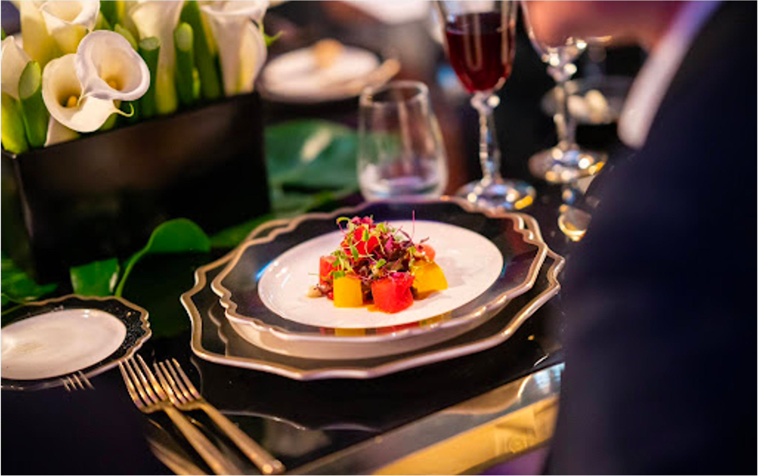 A fine dining table setting with a decorative plate of colorful food, a floral arrangement of white calla lilies in a black vase, and glasses of red wine.