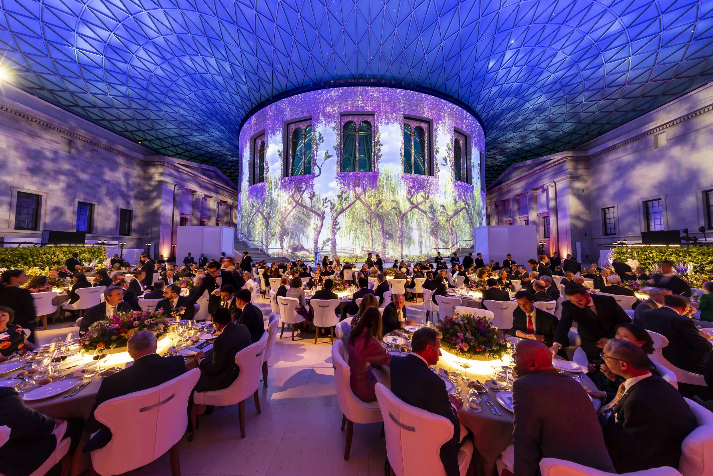 An elegant indoor event space with a high, patterned blue ceiling and large circular projection on the wall depicting a scenic landscape. Attendees are seated at round tables with white chairs, enjoying a formal gathering or dinner.