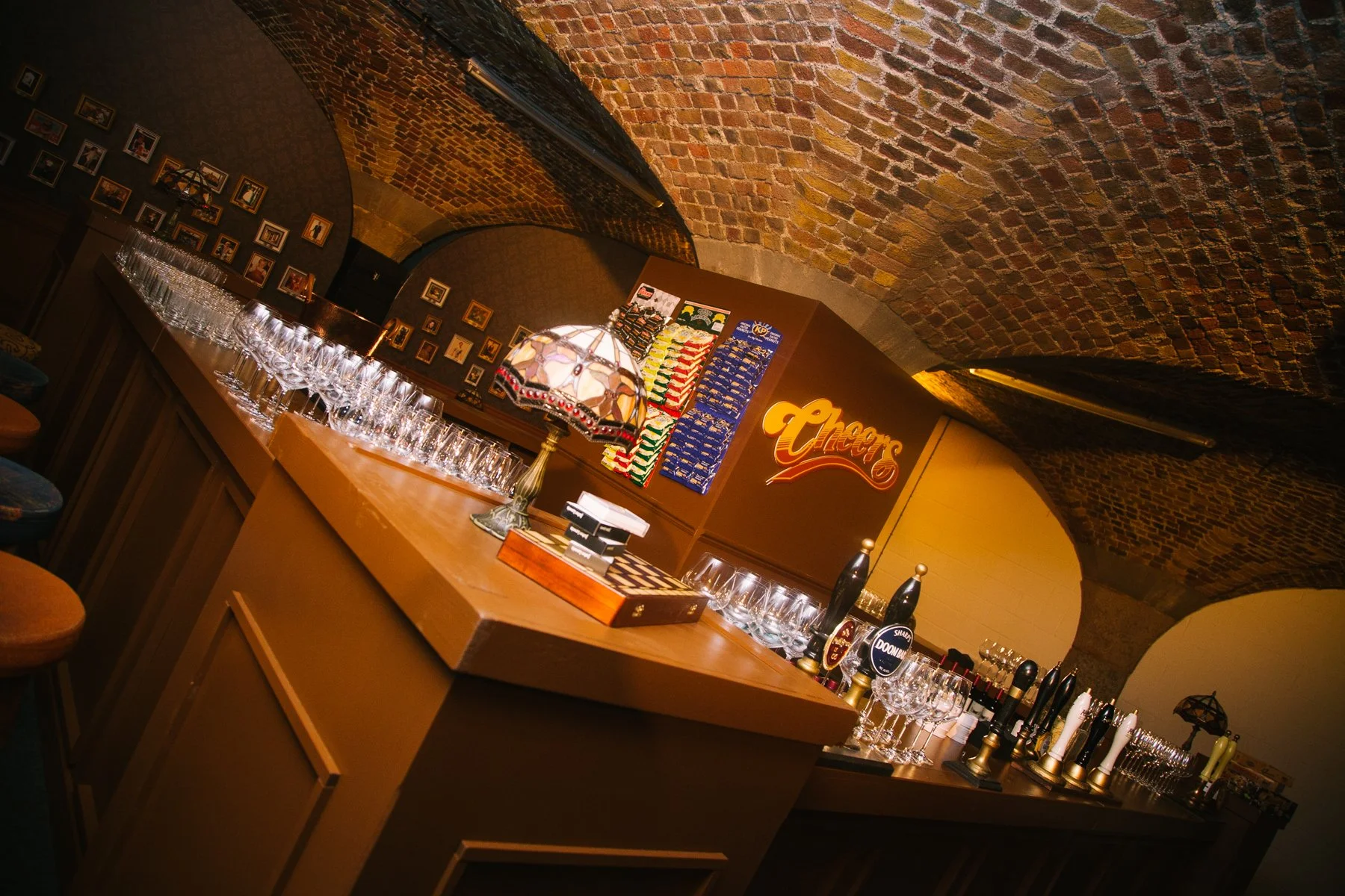 Bar with wooden counter, glassware, beer taps, and decorative lamps, set against brick vaulted ceiling and a wall with framed photos and chalkboard menus.