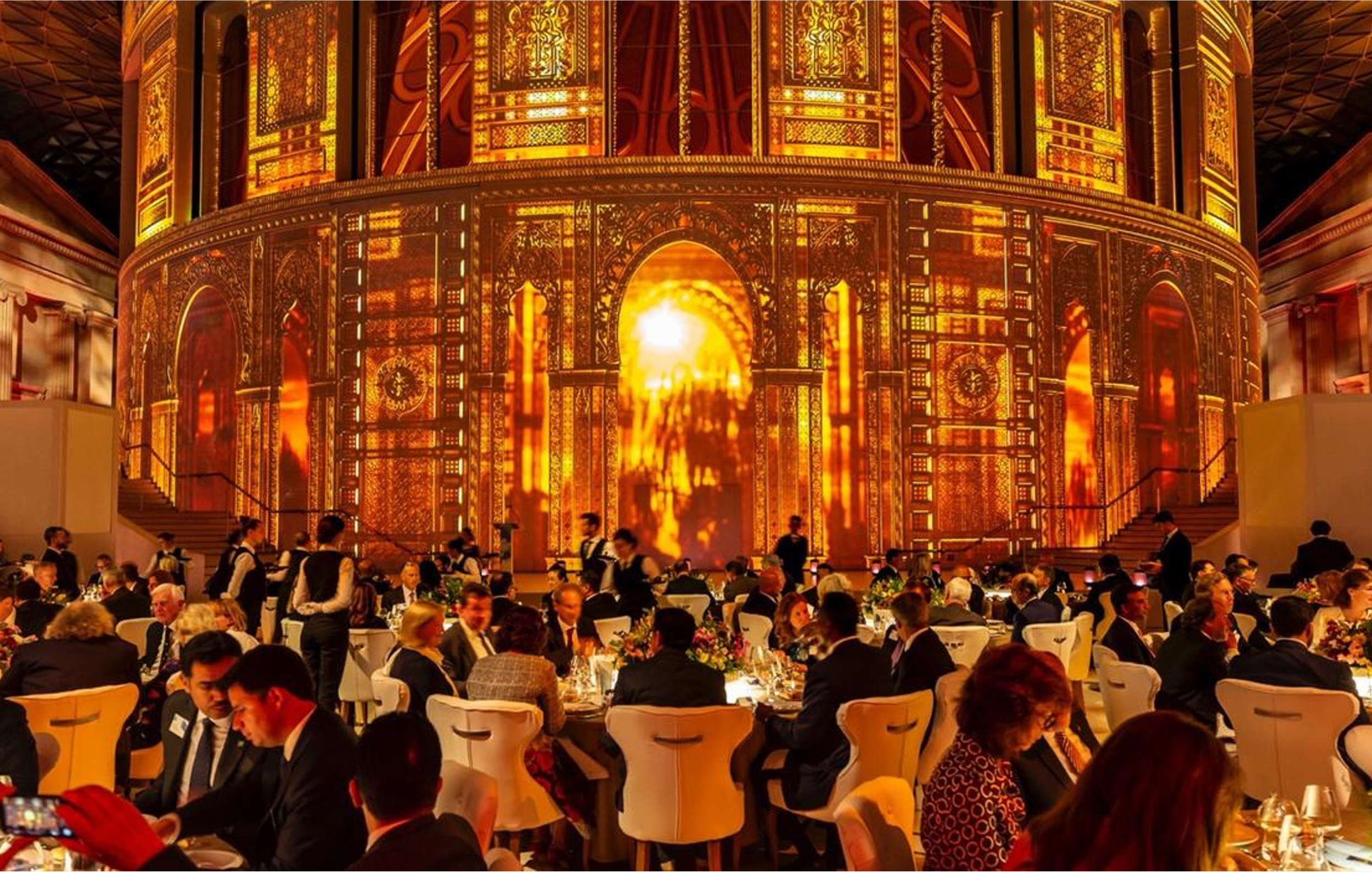 Elegant banquet hall with gold and orange tones, large ornate stage with intricate patterns, and numerous guests seated at round tables enjoying a formal event.