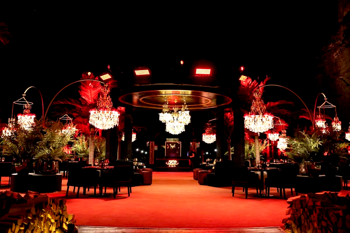 Interior of an upscale lounge or nightclub with red lighting, crystal chandeliers, and black chairs surrounding tables decorated with plants. A stage or DJ booth with the logo 'SASS' is visible in the background.