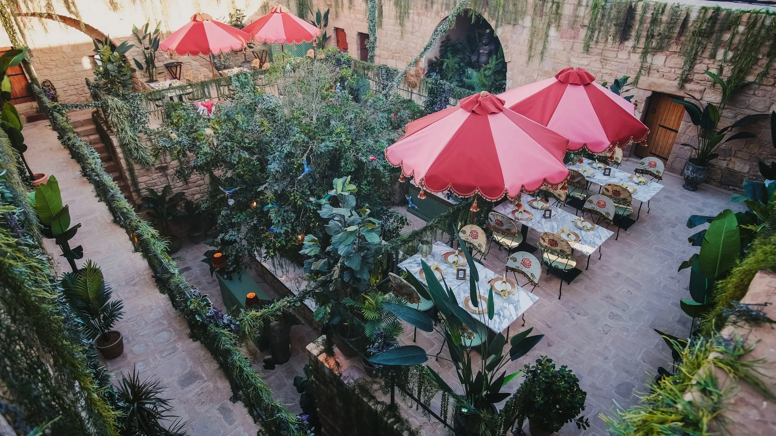 An outdoor dining area with a central tree, surrounded by umbrellas, lush greenery, potted plants, and a stone wall, featuring tables set with dishes and chairs.