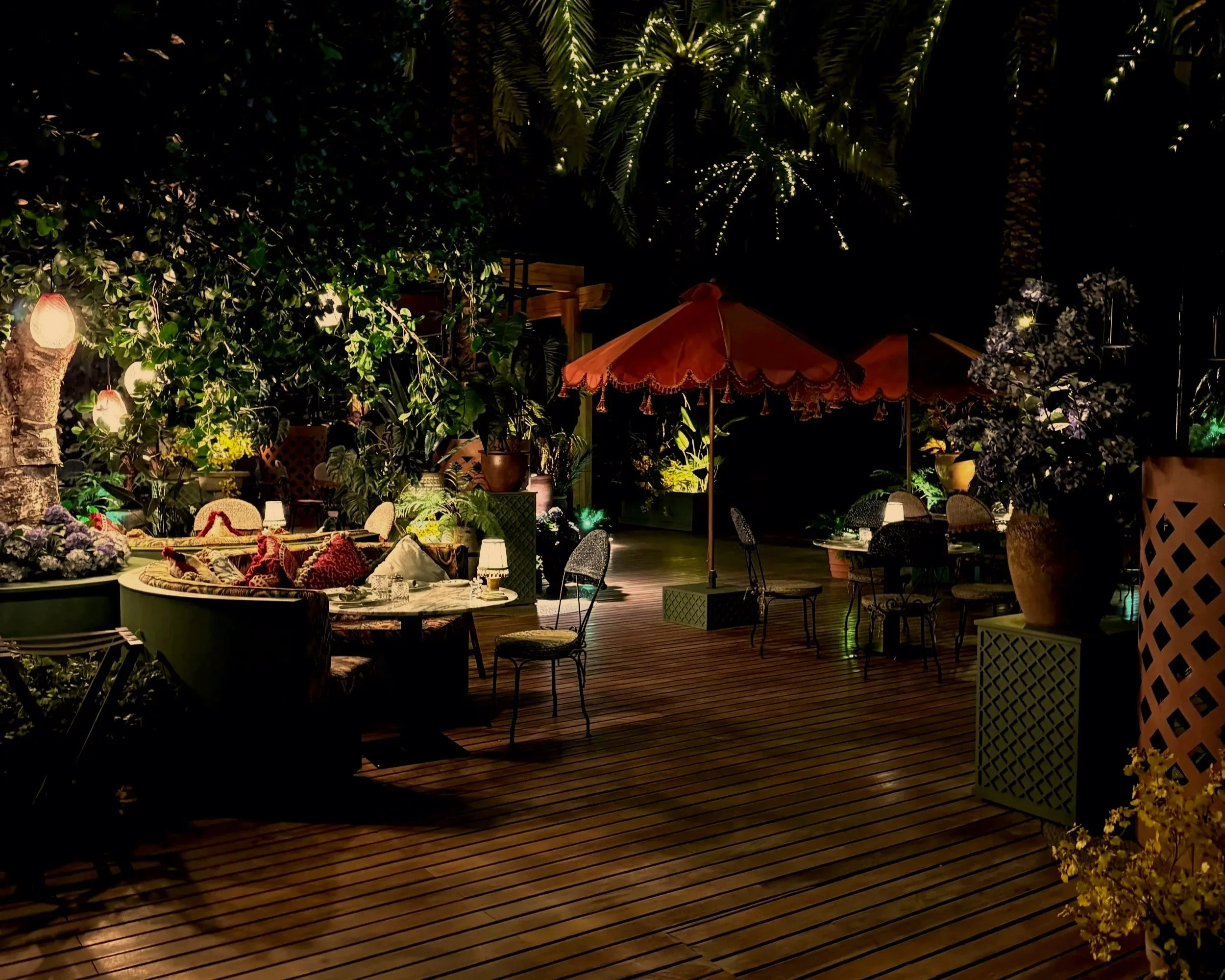 A cozy outdoor patio at night with wooden flooring, lush greenery, string lights, and various seating areas including a table with pillows, a large orange umbrella, and decorative potted plants.