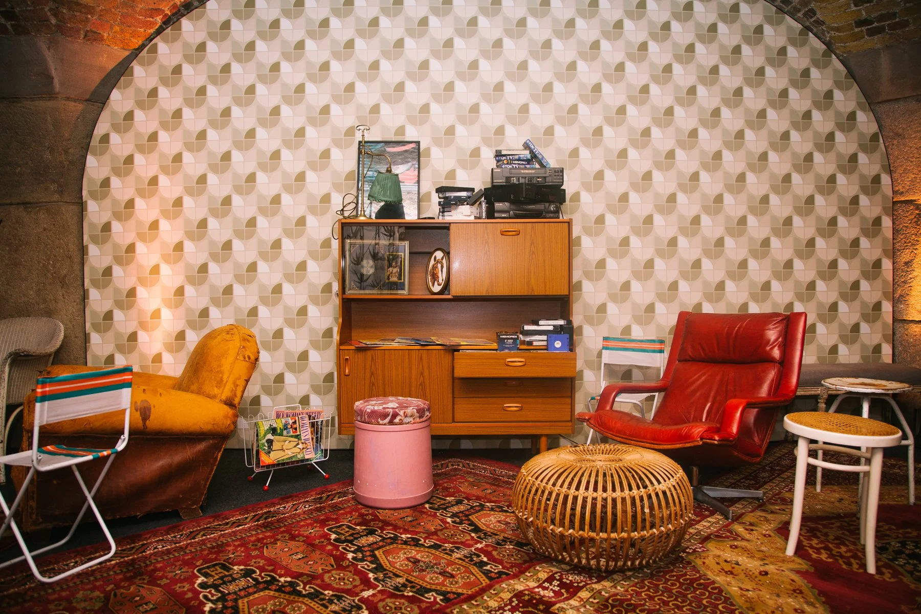 Eclectic vintage living room with patterned wallpaper, red leather armchair, yellow armchair, wicker ottoman, various side tables, a wooden cabinet with magazines and framed photos, and a carpet with intricate designs.