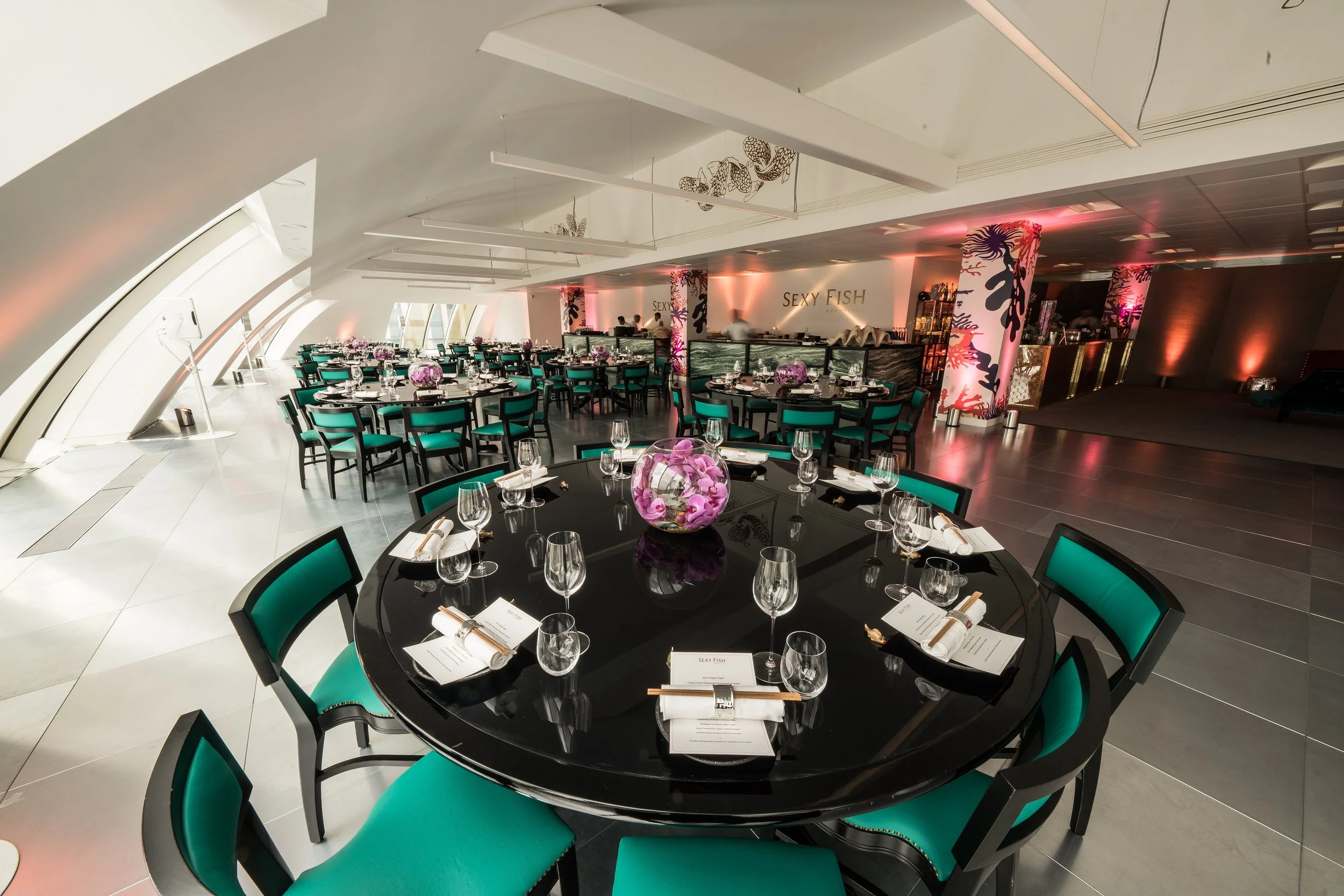 Elegant restaurant with round tables set for dining, decorated with pink floral centerpieces, and teal chairs; modern interior design with arched windows and colorful lighting.