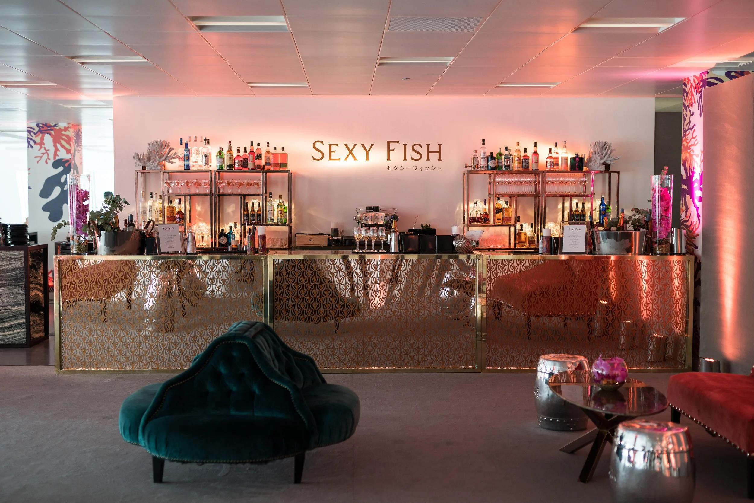 A stylish bar with a gold lattice front, stocked with various bottles of alcohol and glassware, under a sign that reads 'Sexy Fish' with Japanese characters. The bar background has pink lighting, and there's a velvet teal chair and a small table with metallic accents and flowers in the foreground.