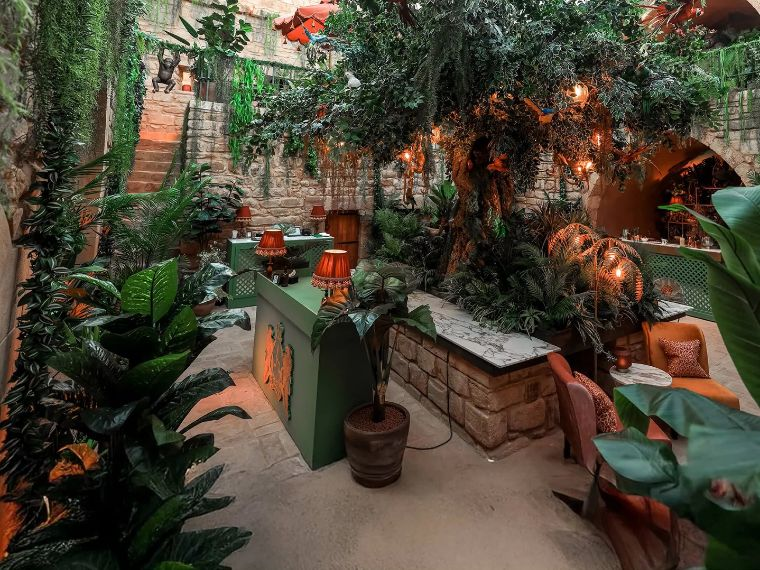 Indoor tropical-themed lounge area with lush greenery, brick walls, and warm lighting, featuring a stone planter and cozy seating.