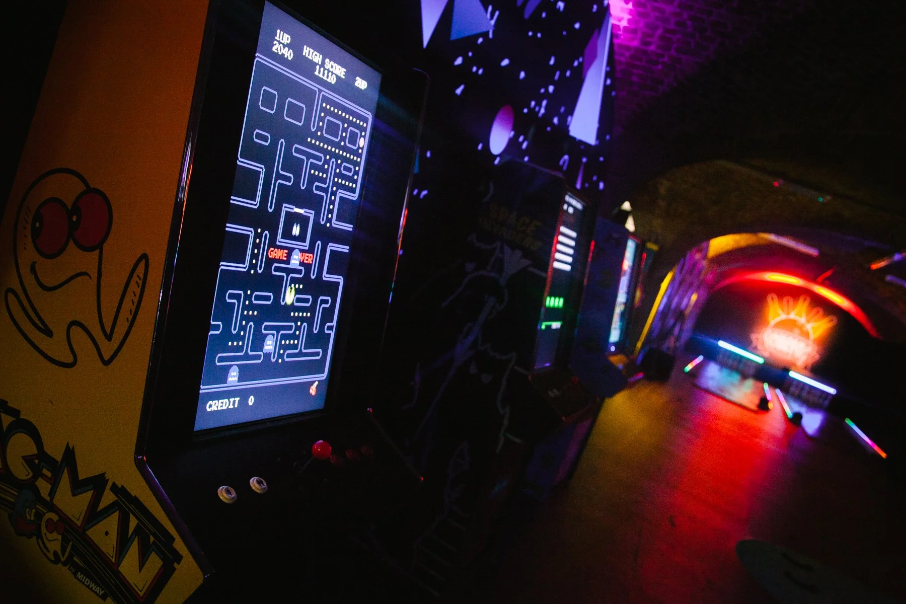 Arcade cabinet playing Pac-Man in a dimly lit arcade with neon lighting and colorful lights in the background.