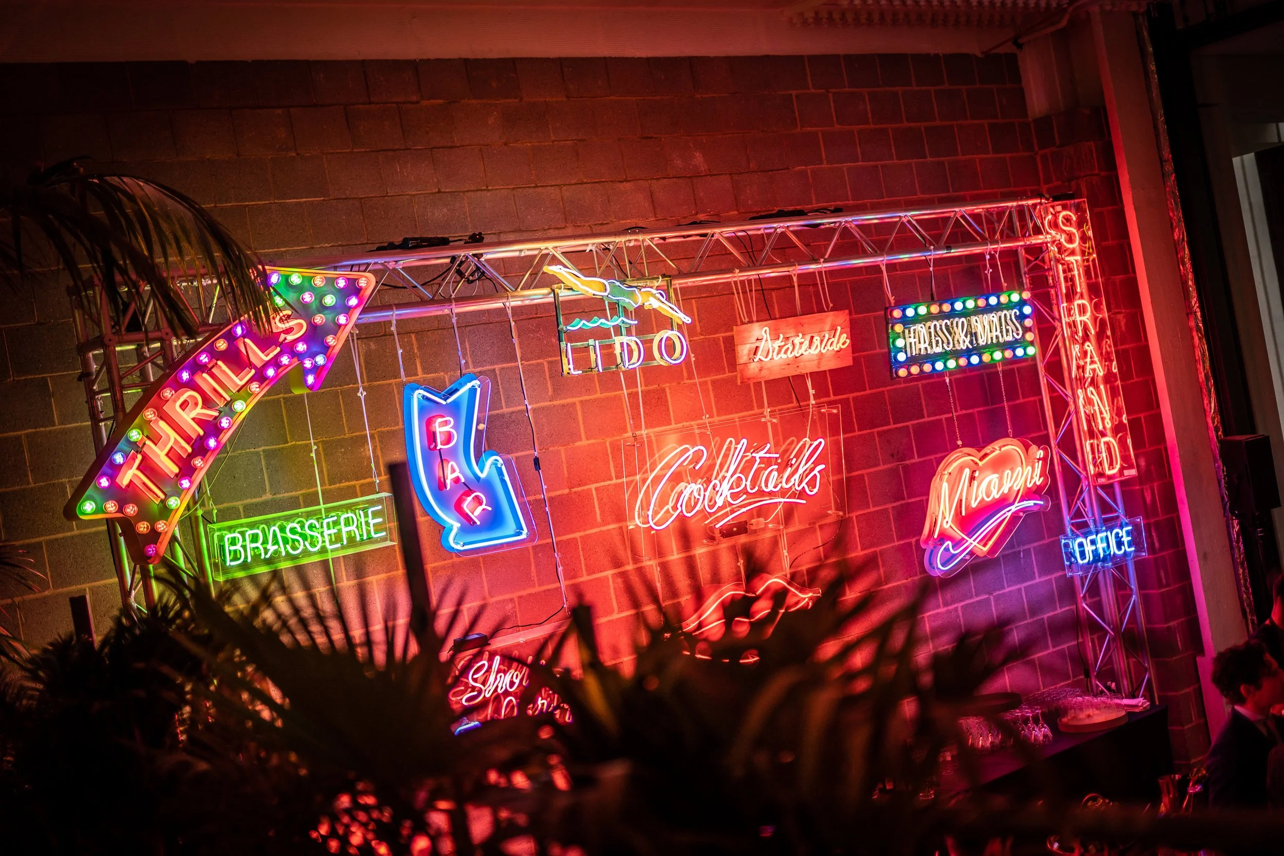 Neon signs on a brick wall advertising a bar with words like 'Tropical,' 'Brasserie,' 'Cocktails,' and 'Miami,' with various colorful lights and decorations.