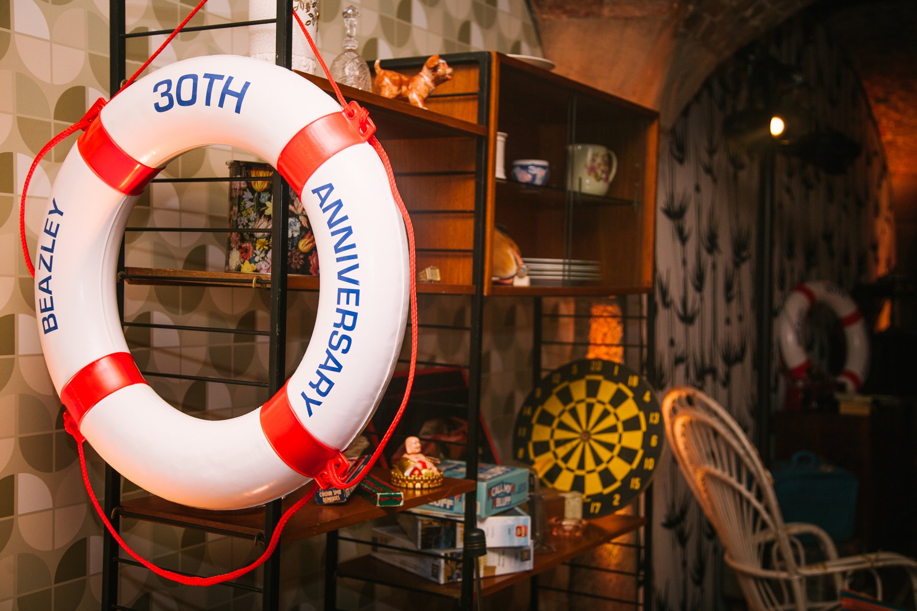 Decorative indoor setting with a lifebuoy that has 'BEAZLEY 30TH ANNIVERSARY' written on it hanging on a black metal shelf. The shelf also holds figurines, dishes, and a box, with a dartboard visible in the background along with a white wicker chair and patterned wallpaper.