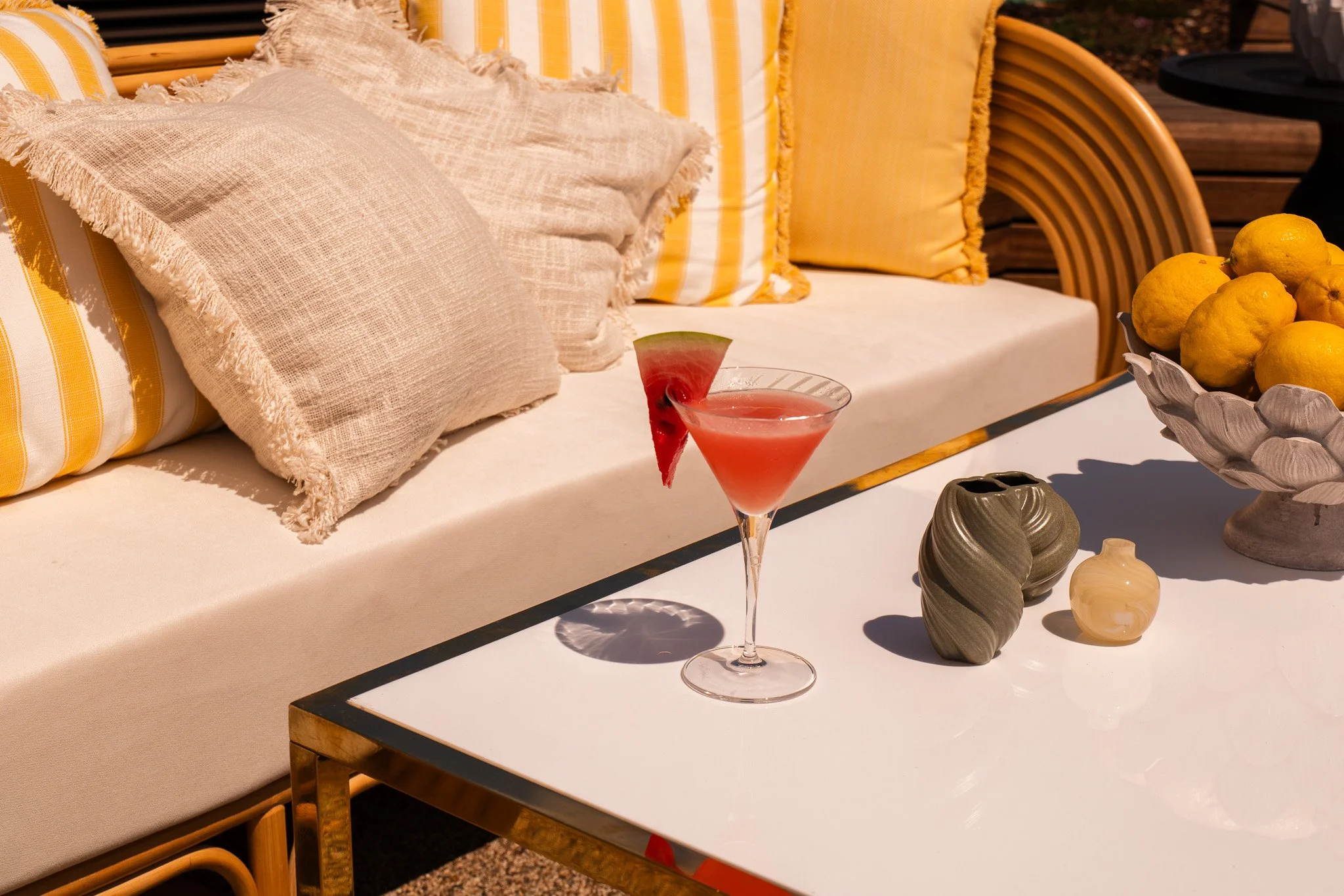 A pink cocktail with a watermelon slice garnish on a white table. The table also holds a ceramic bowl of lemons and two decorative vases. Behind the table, there is a cushioned outdoor sofa with striped yellow and white pillows.